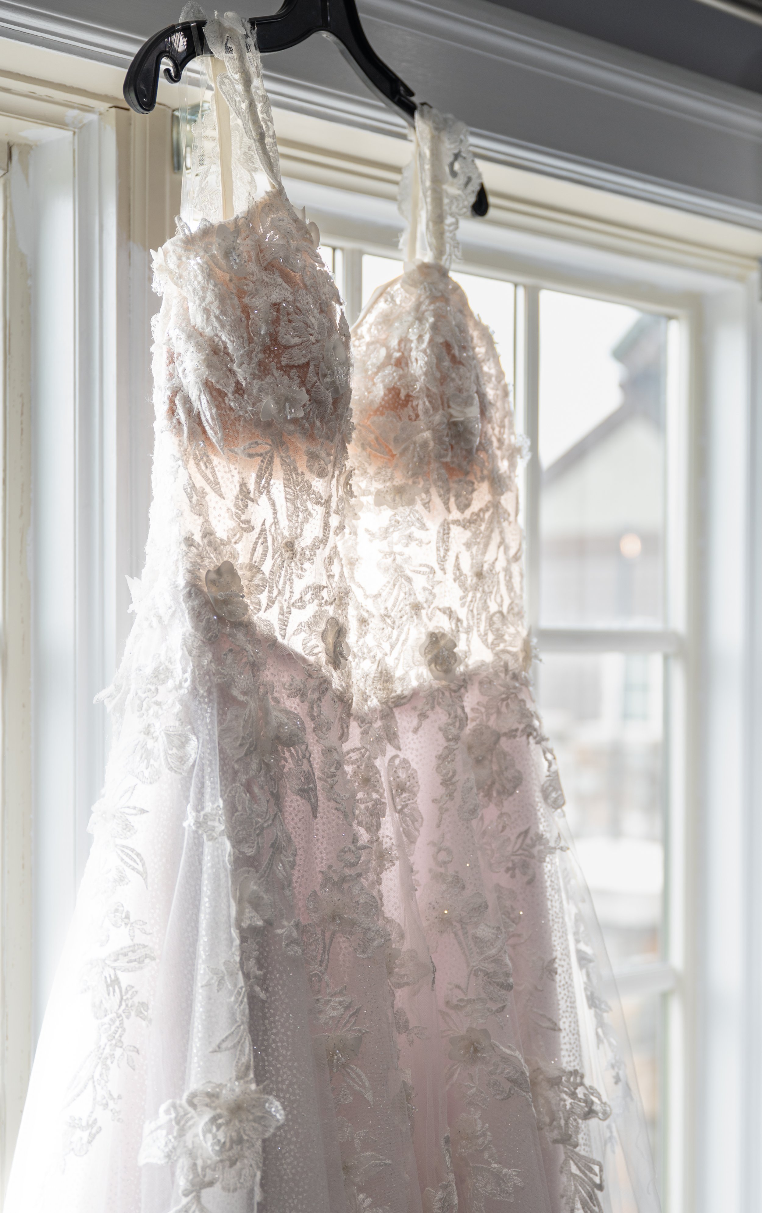 A wedding dress with lace and floral embroidery hanging on a black hanger in front of a window.