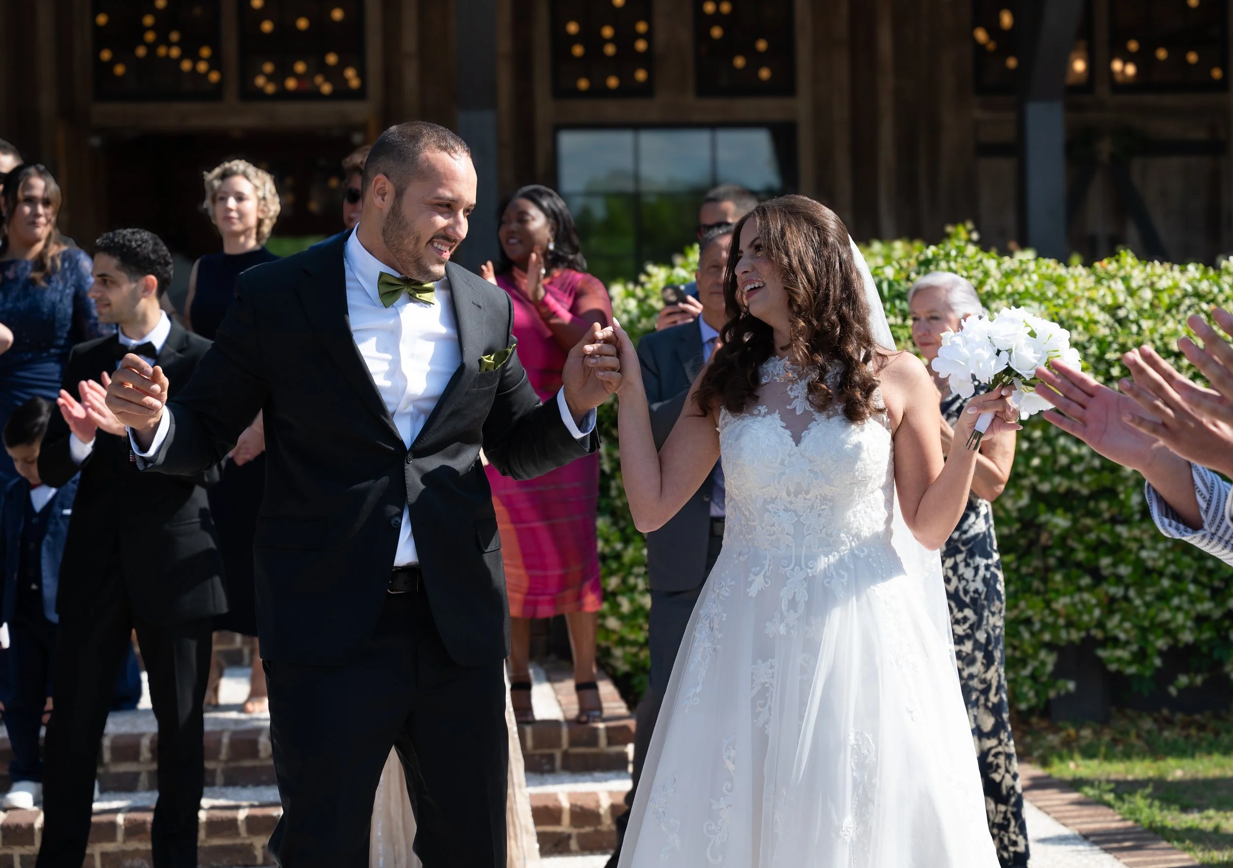 A newlywed couple dancing outdoors at their wedding ceremony, with friends and family clapping and celebrating around them.