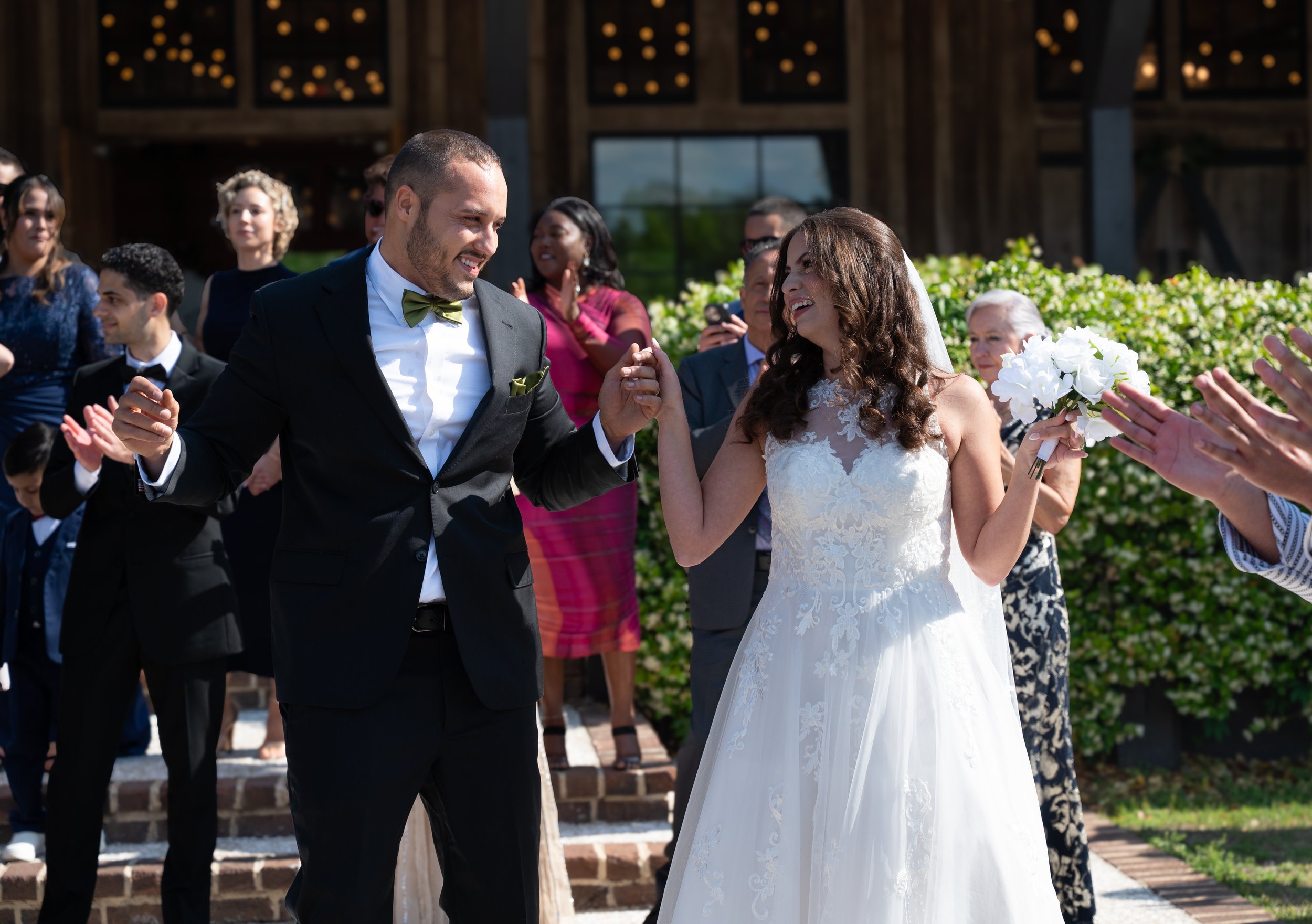 A bride and groom celebrating their wedding outdoors, holding hands and smiling at each other, surrounded by friends and family.
