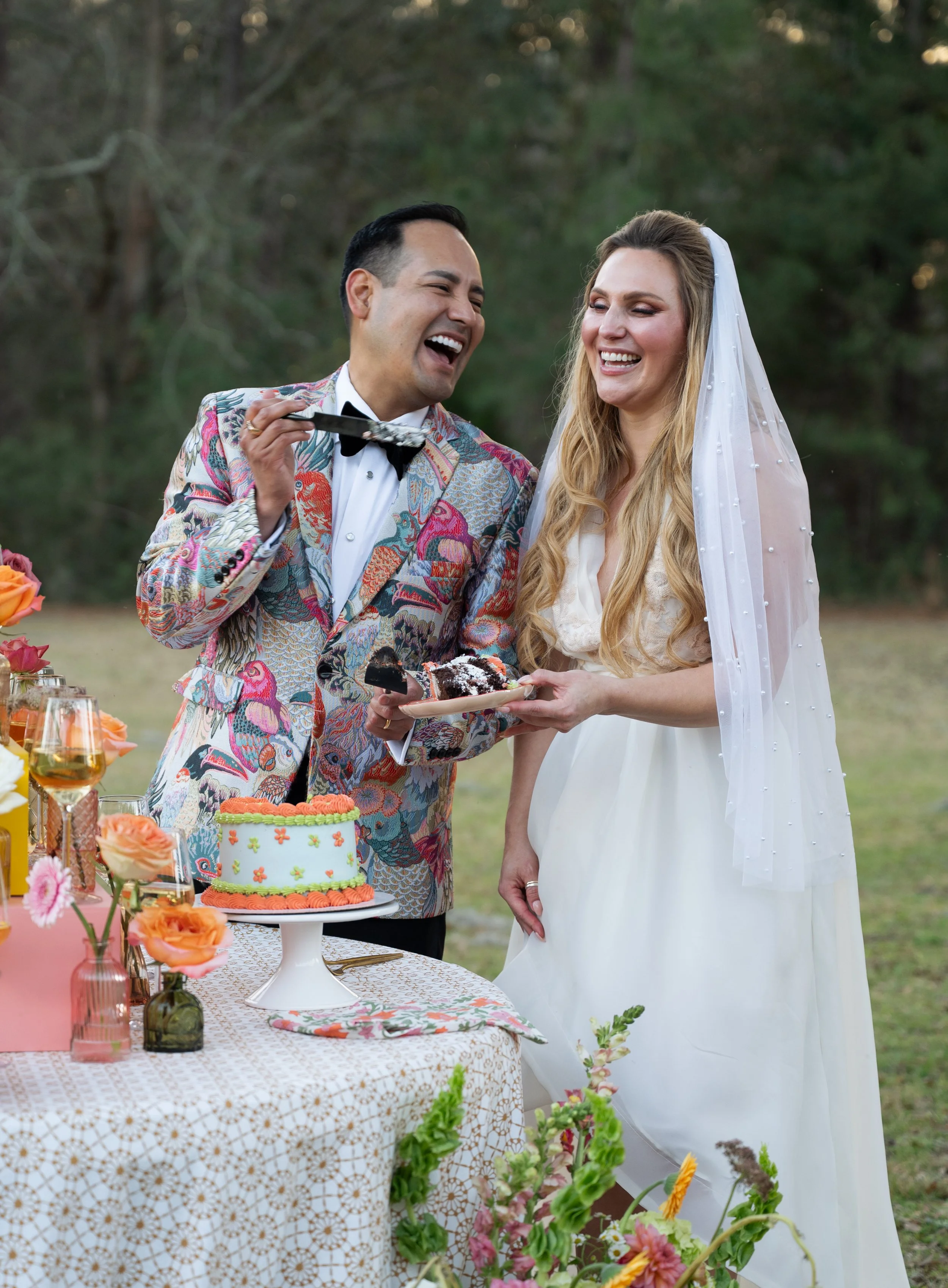 A newlywed couple celebrating at an outdoor wedding reception. The groom is wearing a colorful, patterned suit and is laughing while holding a piece of cake. The bride, in a white dress with a veil, is smiling while holding a plate with cake. There i