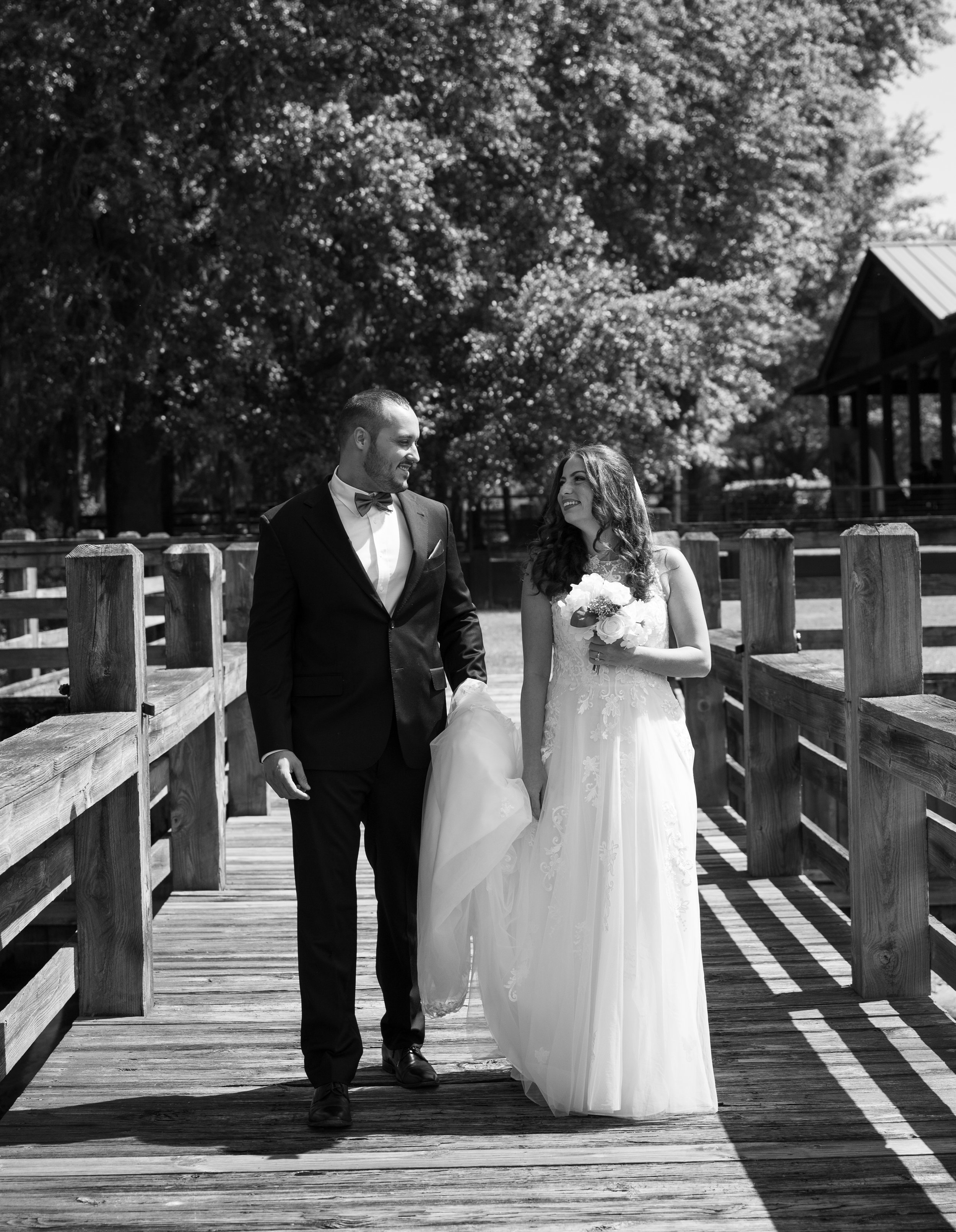 A man in a tuxedo and bow tie and a woman in a wedding dress holding a bouquet walk together on a wooden bridge outdoors, smiling at each other with trees and a building in the background.