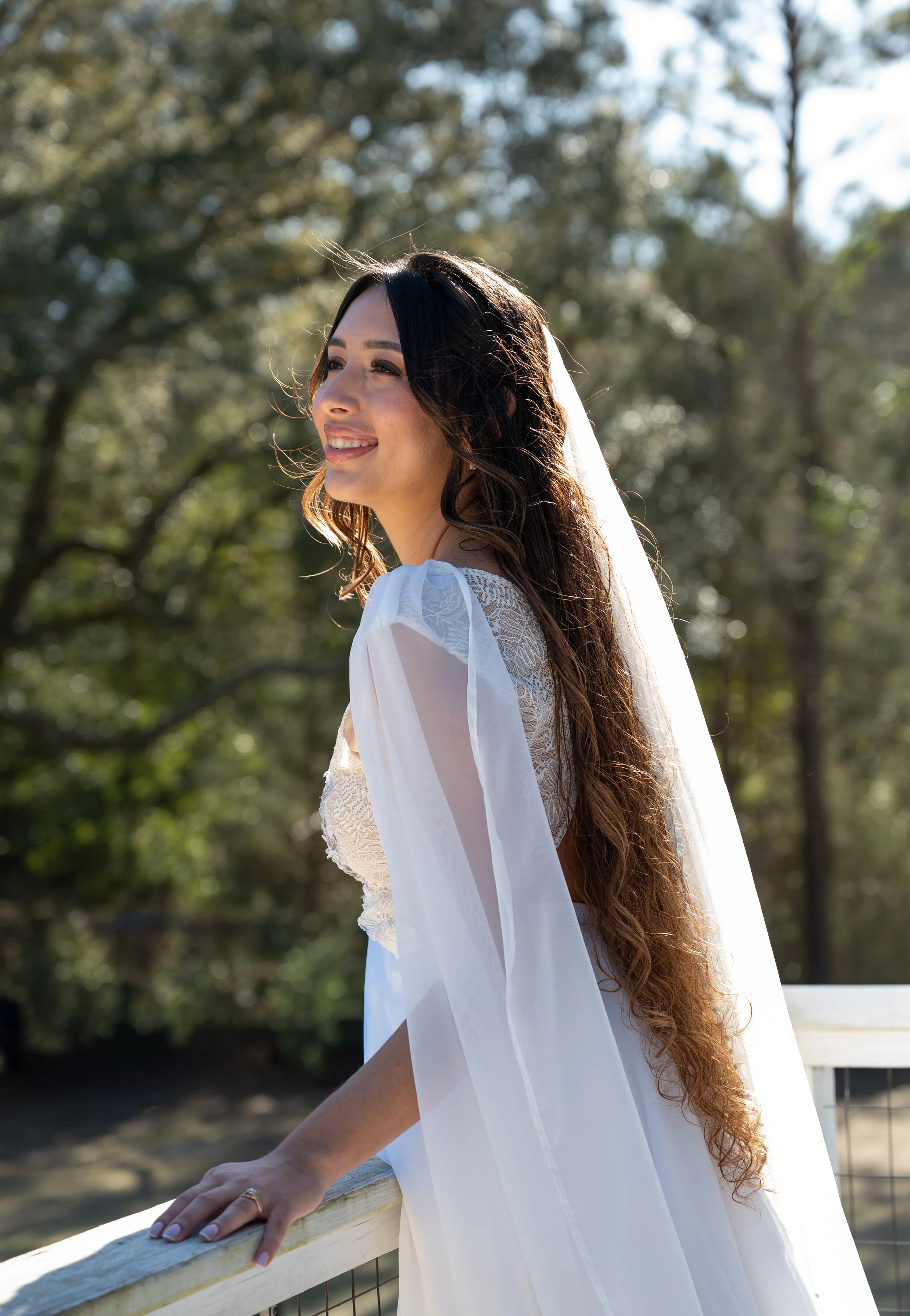 A woman with long dark hair and a wedding veil, dressed in a white lace dress, standing outdoors on a sunny day near a wooden railing, with trees in the background.