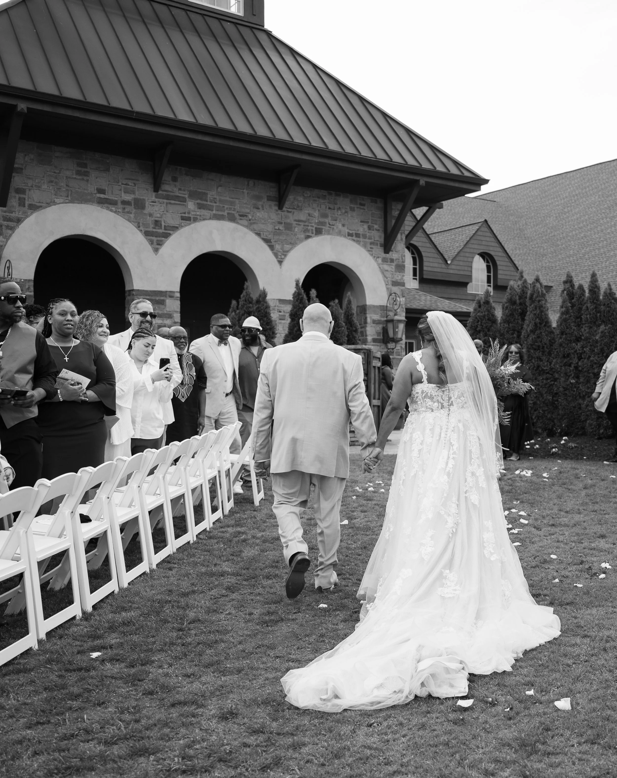 A bride and groom holding hands and walking towards an outdoor wedding ceremony, with guests standing and watching in front of a brick building with an arched entrance.
