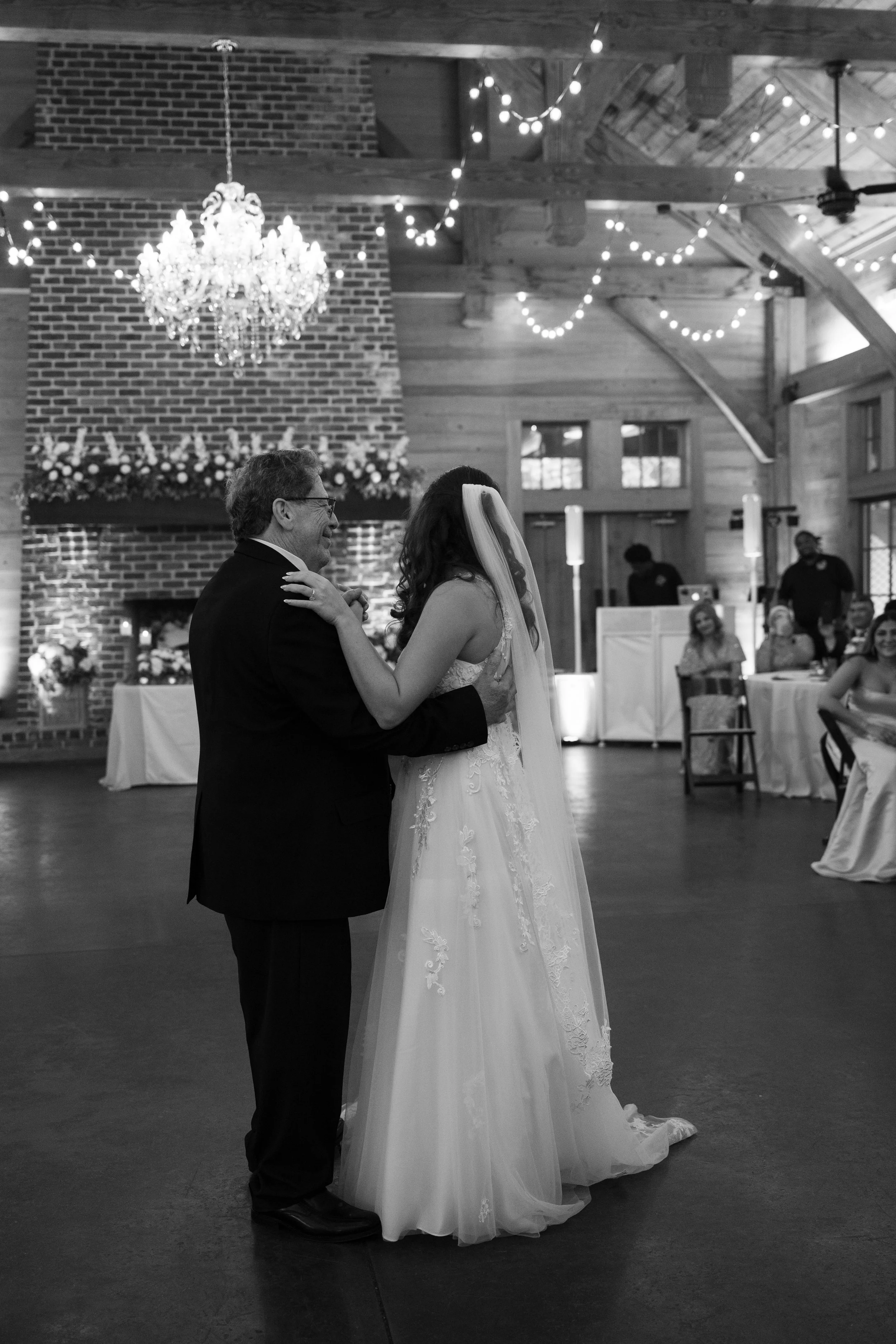 A bride and an elderly man share a father-daughter dance at a wedding reception in a rustic venue with a brick fireplace, decorated with a chandelier and string lights.
