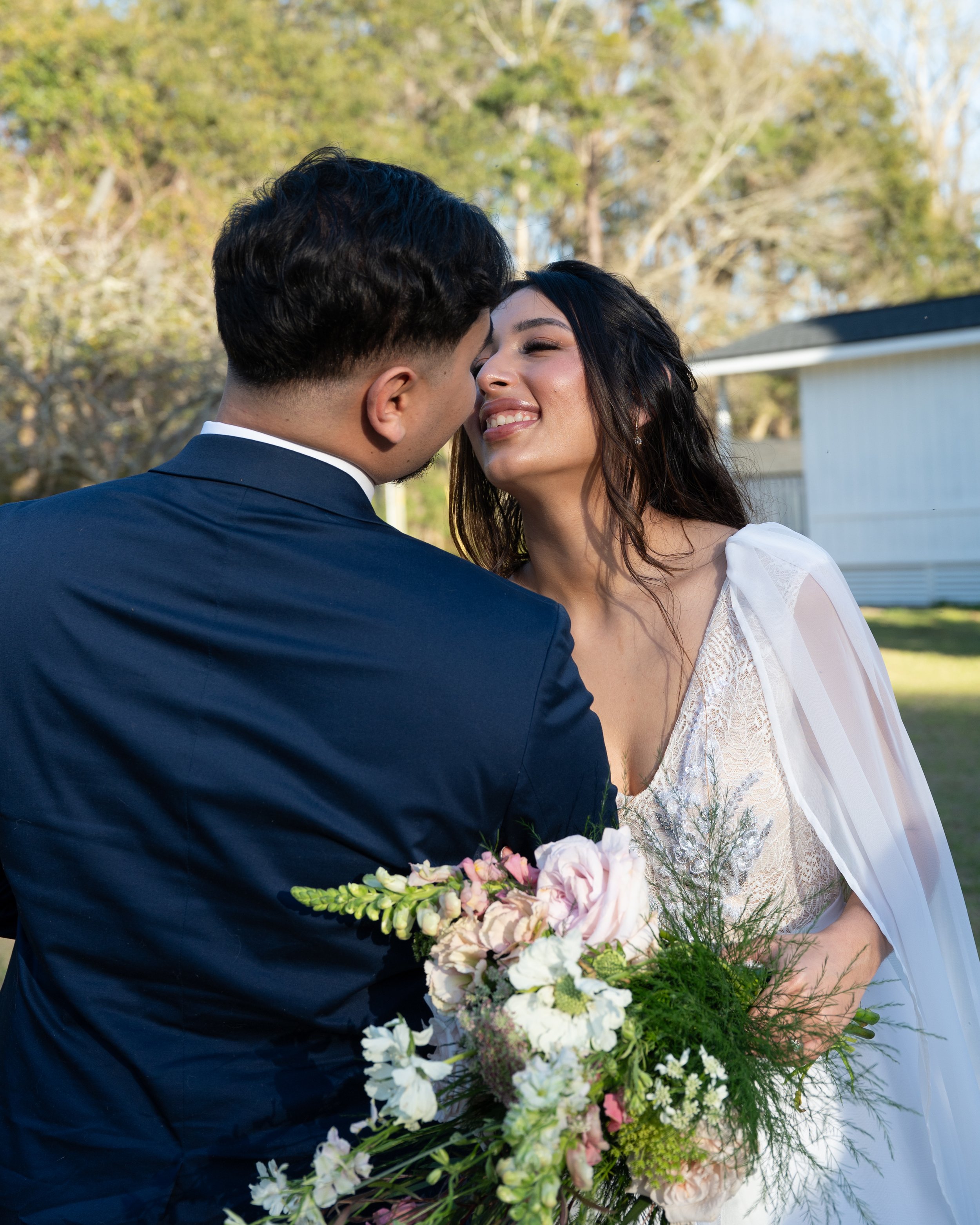 A couple dressed in wedding attire, with the groom in a navy suit and the bride in a white dress, sharing a close moment outdoors with trees in the background and holding a large bouquet of flowers.