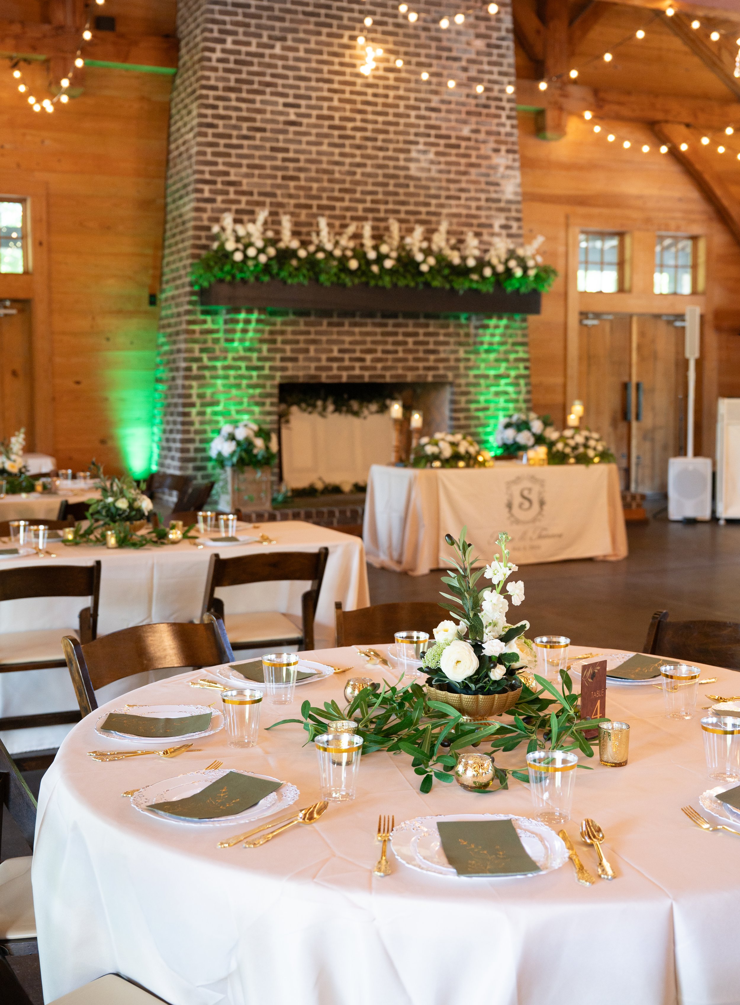 A round banquet table decorated with a white tablecloth, gold flatware, green and gold chargers, and grey napkins. A floral centerpiece with white flowers and greenery sits in a gold basket in the center of the table. Small gold votive candles are sc