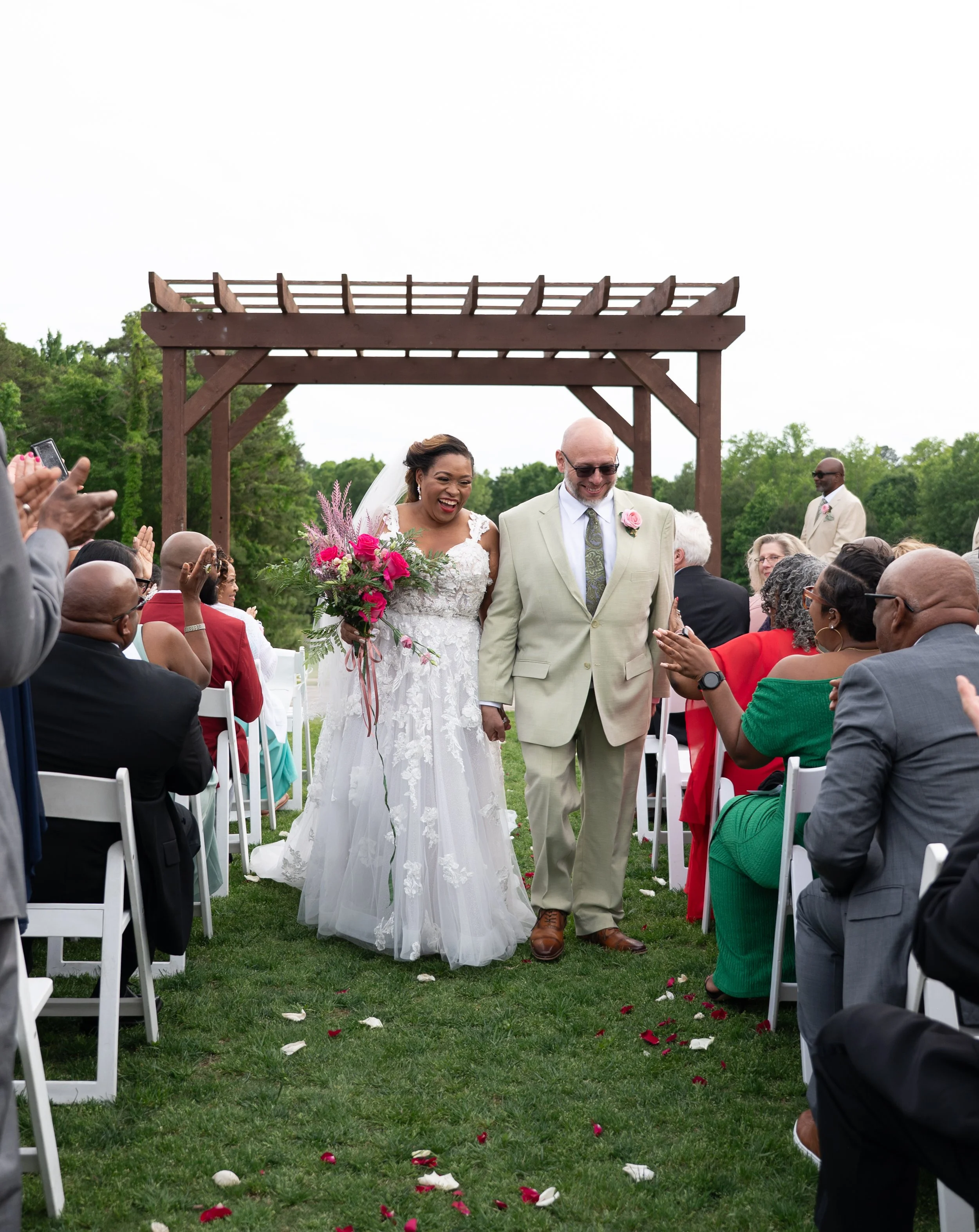 A bride and an older groom walking down the aisle at an outdoor wedding ceremony, surrounded by seated guests, with a wooden arch and greenery in the background.