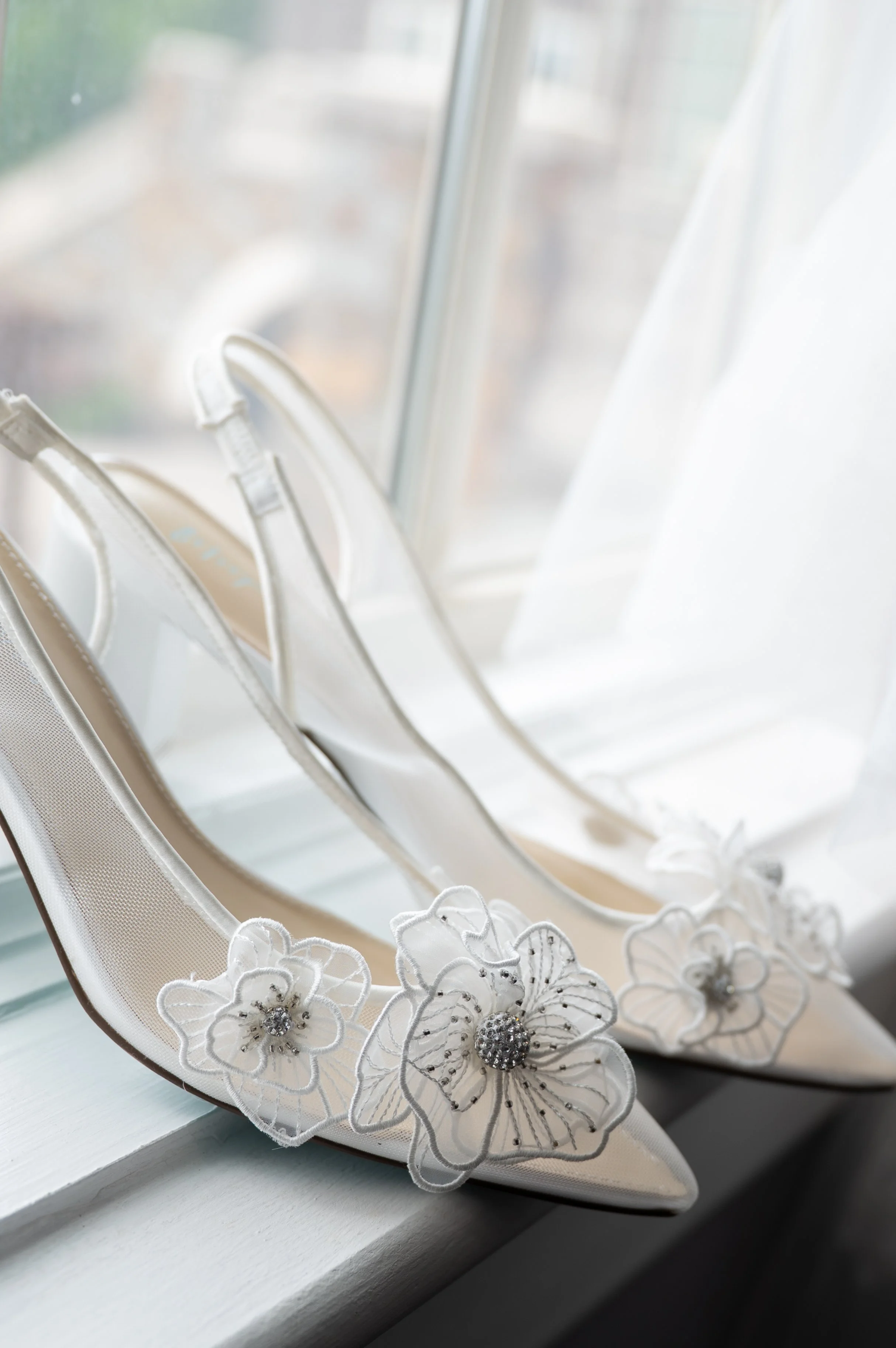 White high-heeled shoe with floral embellishments and rhinestones, placed on a windowsill with lace detailing at the back, viewed from the side.