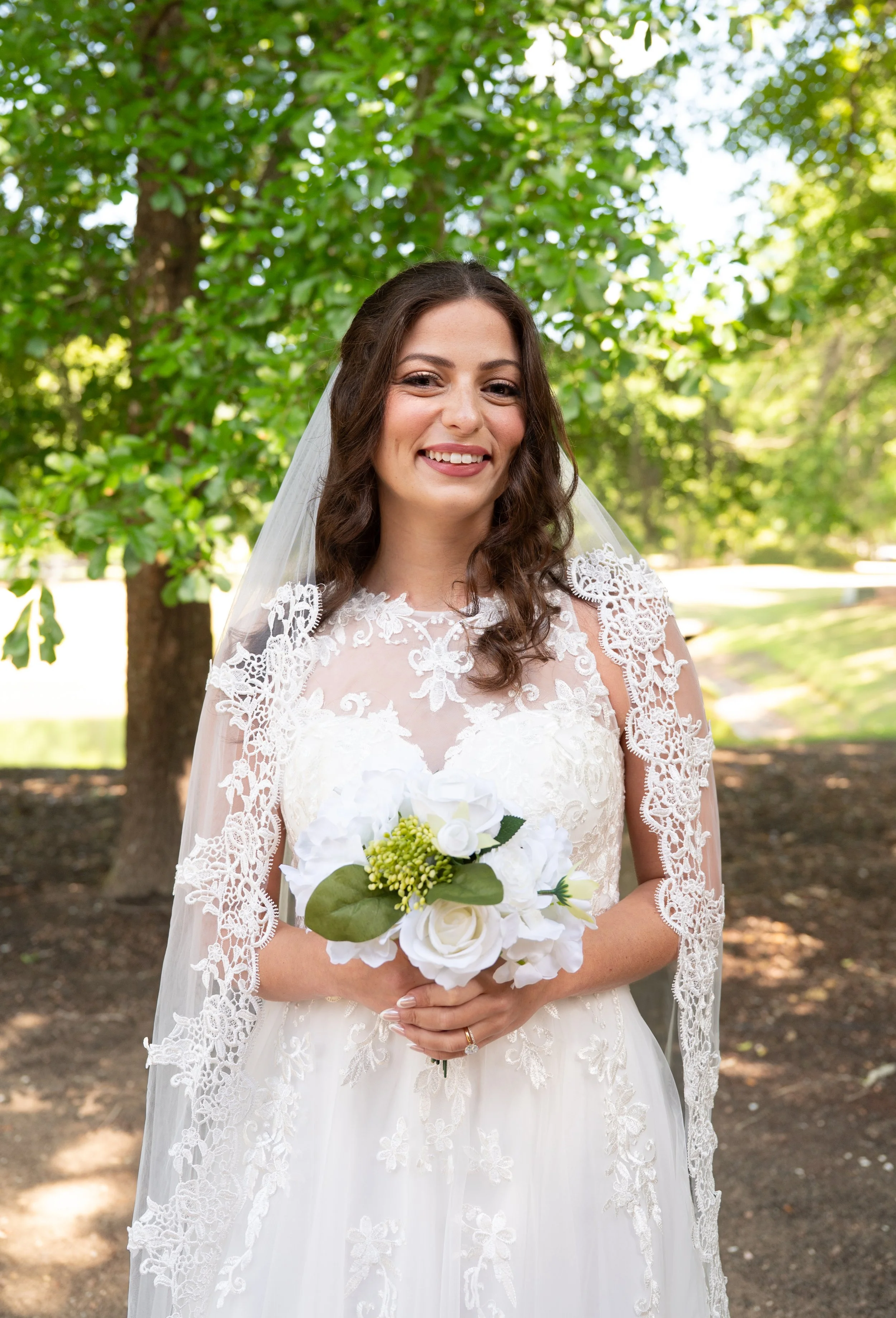 A smiling woman in a wedding dress holding a bouquet of white flowers, standing outdoors in front of green trees.