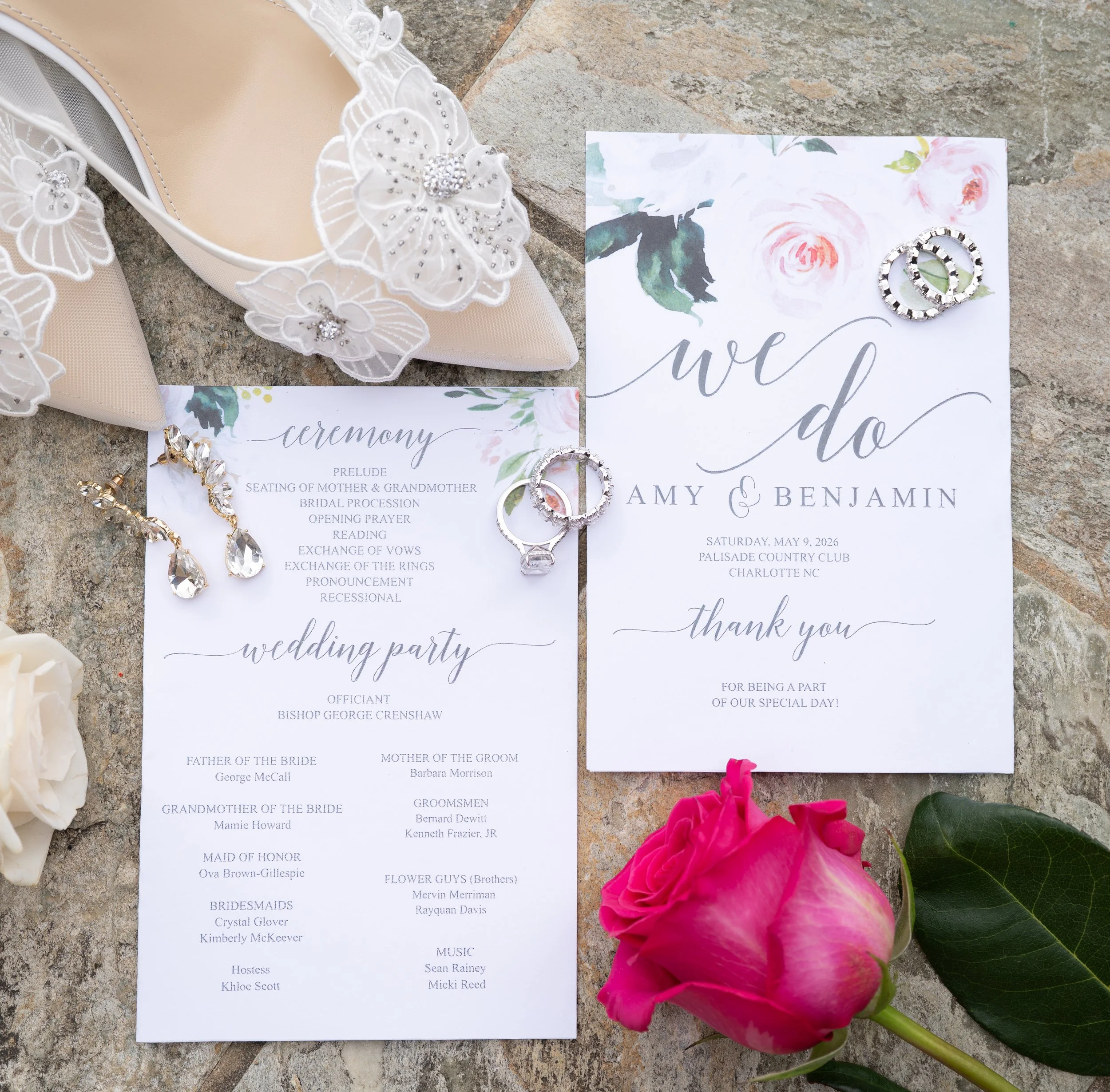Wedding ceremony program on a stone surface surrounded by pink and white flowers and decorative rings.