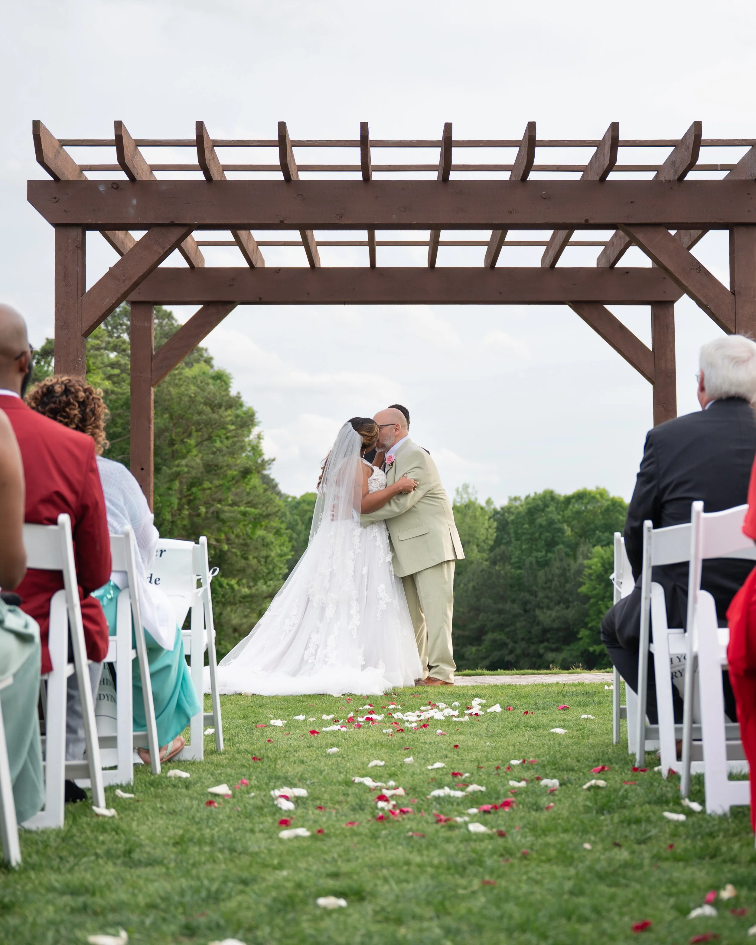 A wedding ceremony taking place outdoors with a couple kissing under a wooden arch, surrounded by seated guests on grassy ground with flower petals scattered.