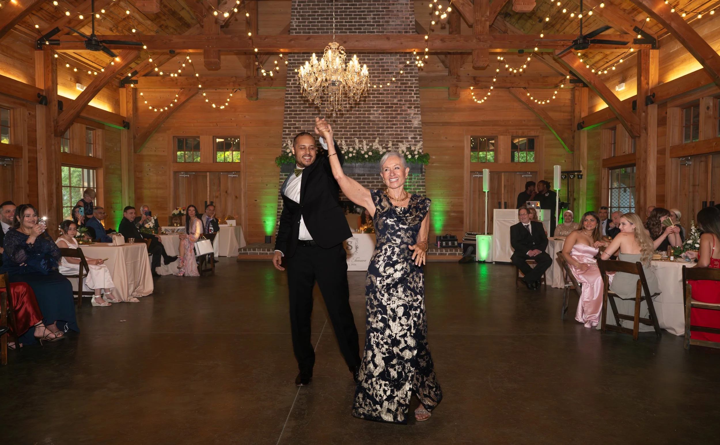 Two people, a man in a tuxedo and a woman in a fancy dress, are dancing and smiling at a wedding reception. The background features a decorated wooden hall with string lights, a chandelier, and guests seated at decorated tables.