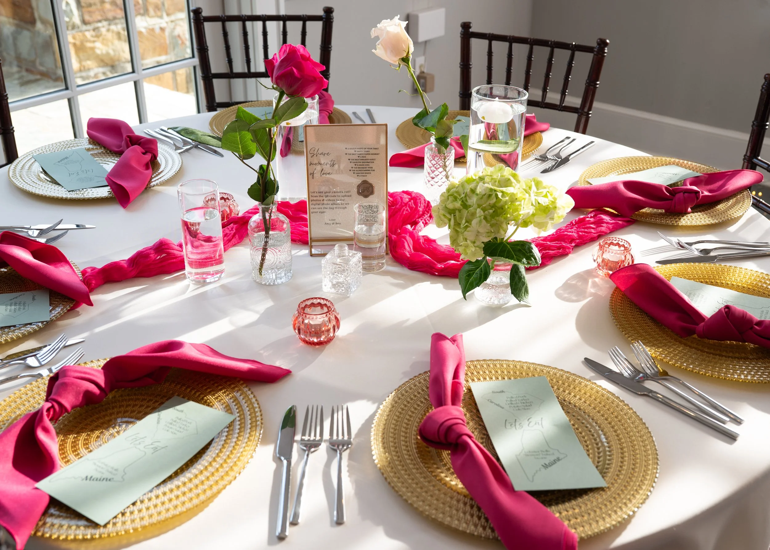 A round dining table decorated for a special occasion with gold placemats, pink napkins tied in a knot, floral centerpieces with pink and white roses, and small pink votive candles.