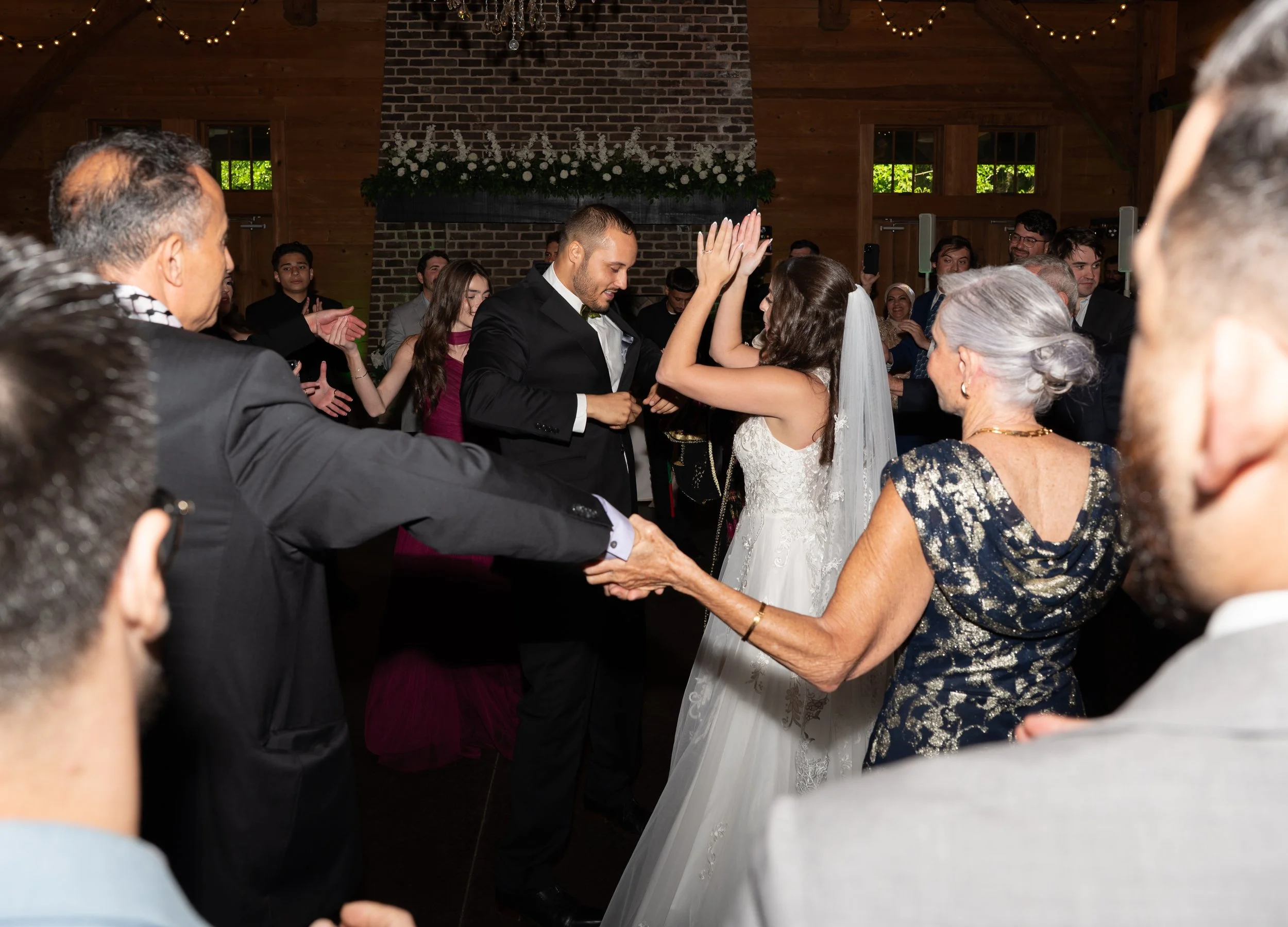 A bride and groom dancing at their wedding reception with guests surrounding them in a rustic venue decorated with string lights and flowers.