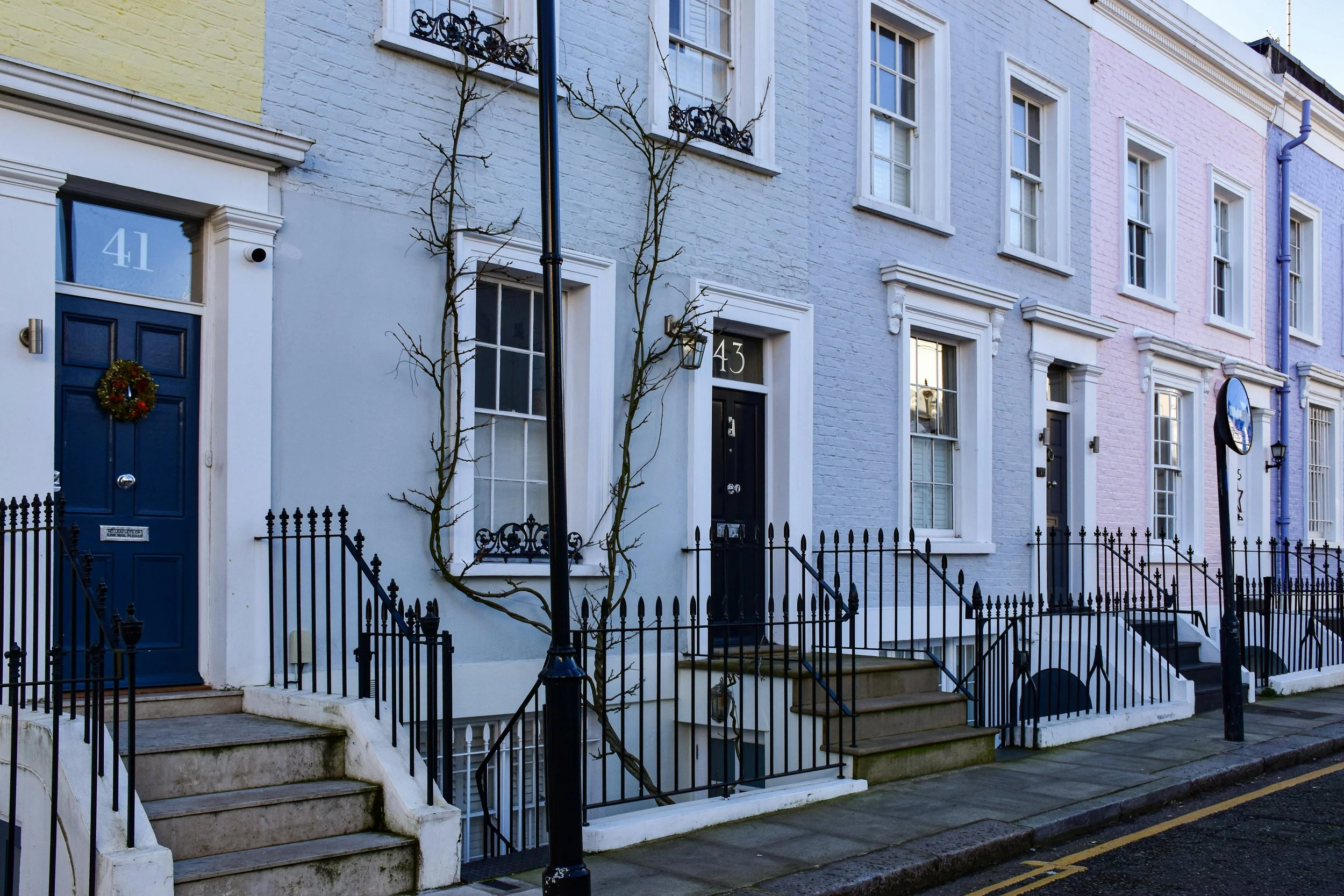 Row of colorful terraced houses with stairs, black railings, and street lamps, featuring doors numbered 41 and 43, some with wreaths and bare trees in front.