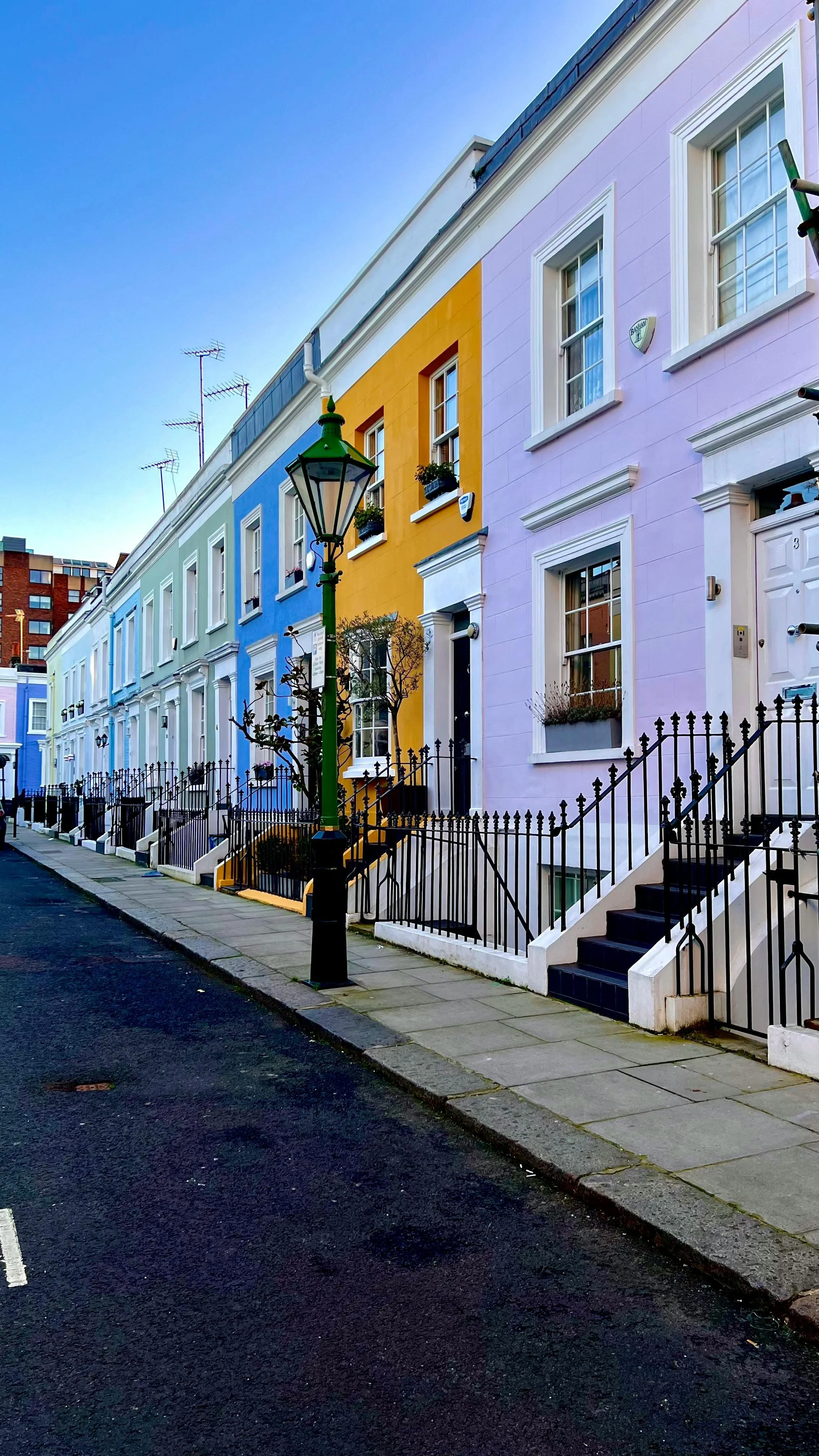 Colorful row of houses painted in pastel shades of blue, yellow, and pink along a city street with black railings, stairs, and a lamppost.