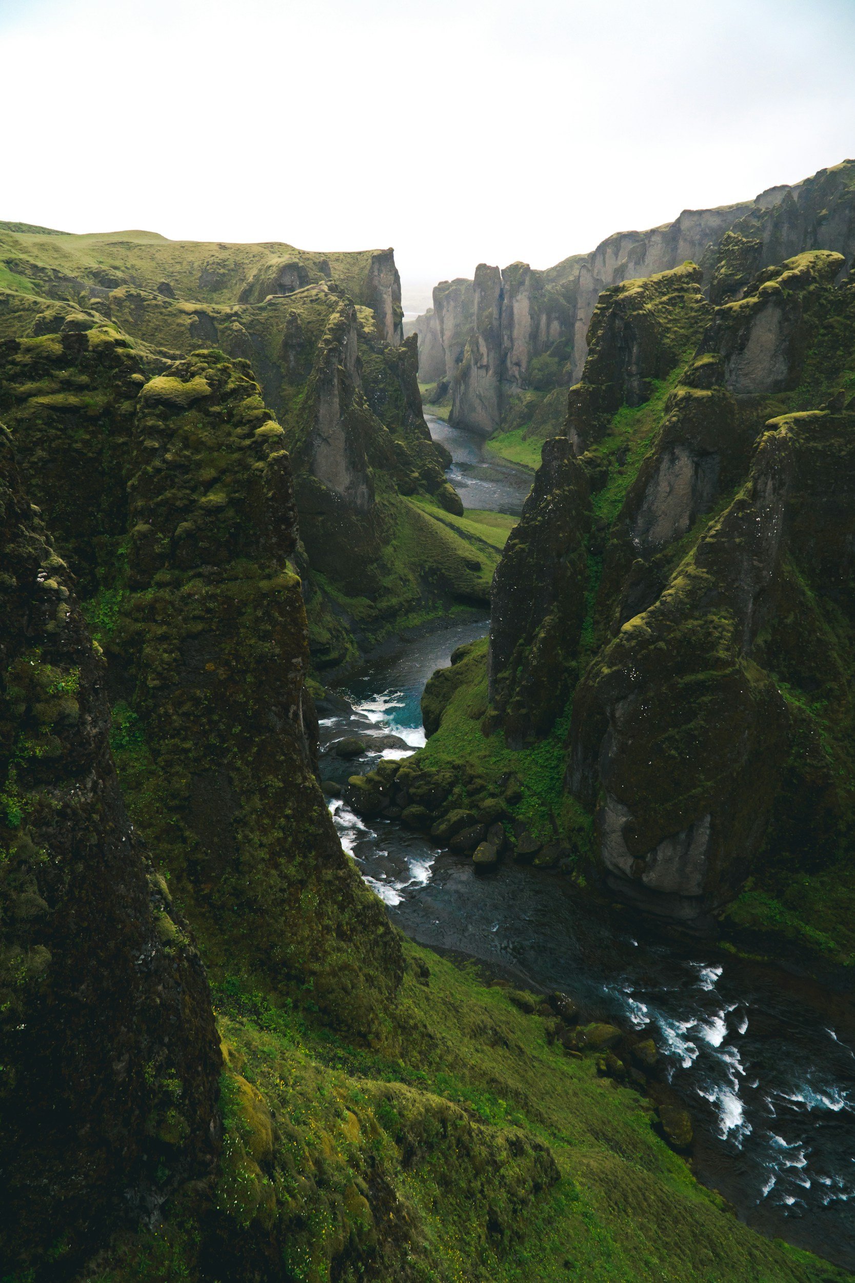 A lush green canyon with steep moss-covered cliffs and a winding river flowing through the bottom.