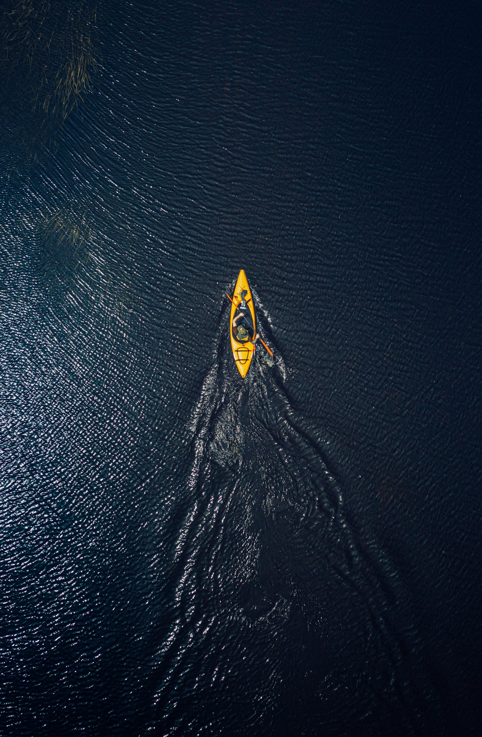 An aerial view of a person kayaking on dark water, paddling forward creating ripples.