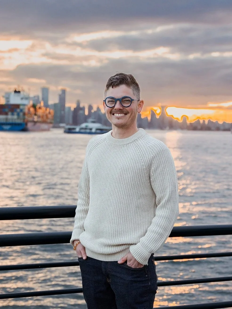 Man with glasses smiling at sunset by the water with city skyline in the background.