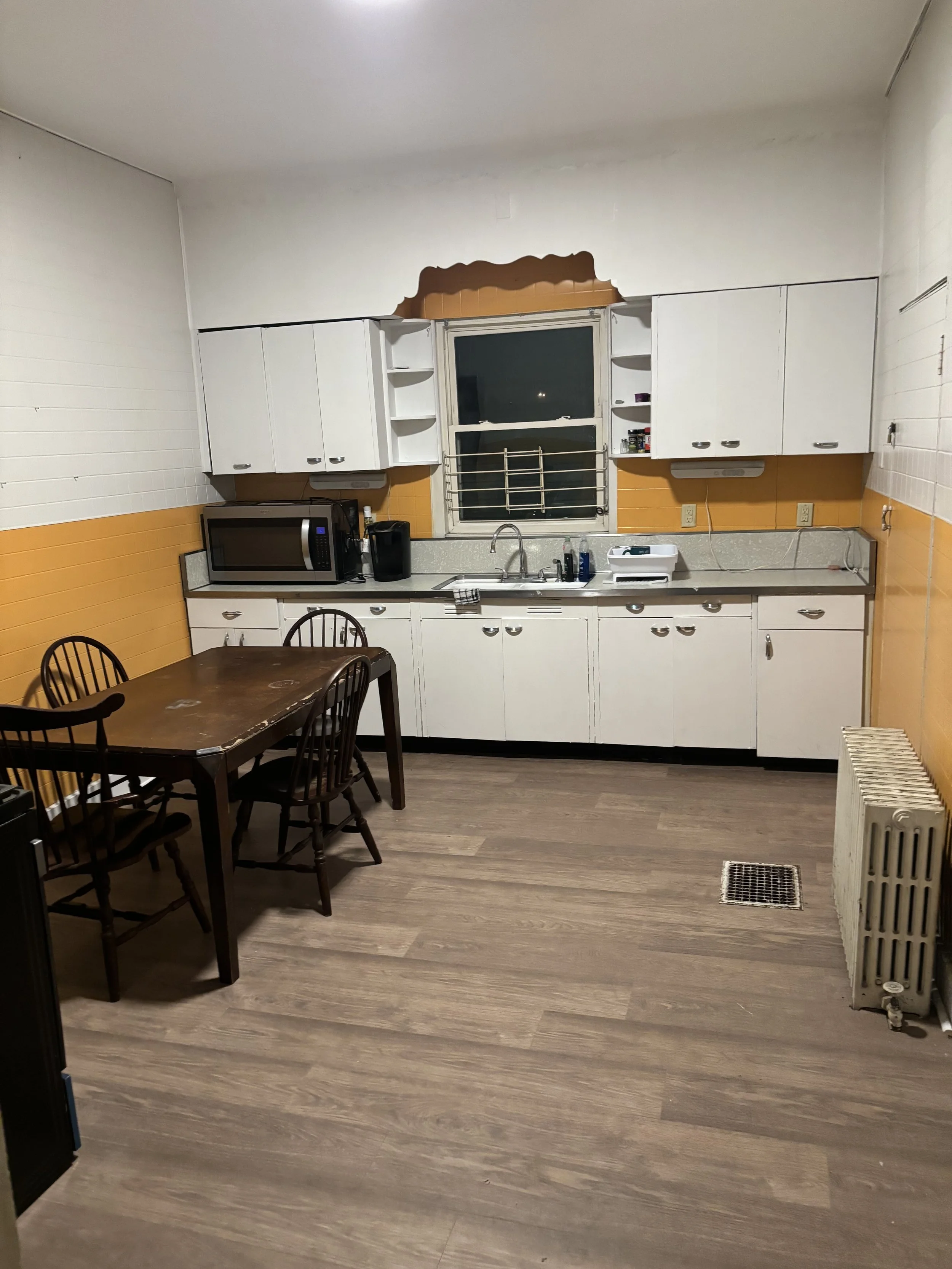 Empty kitchen with white cabinets, orange wall accents, a window with louvers, microwave, dining table with four chairs, and a radiator.