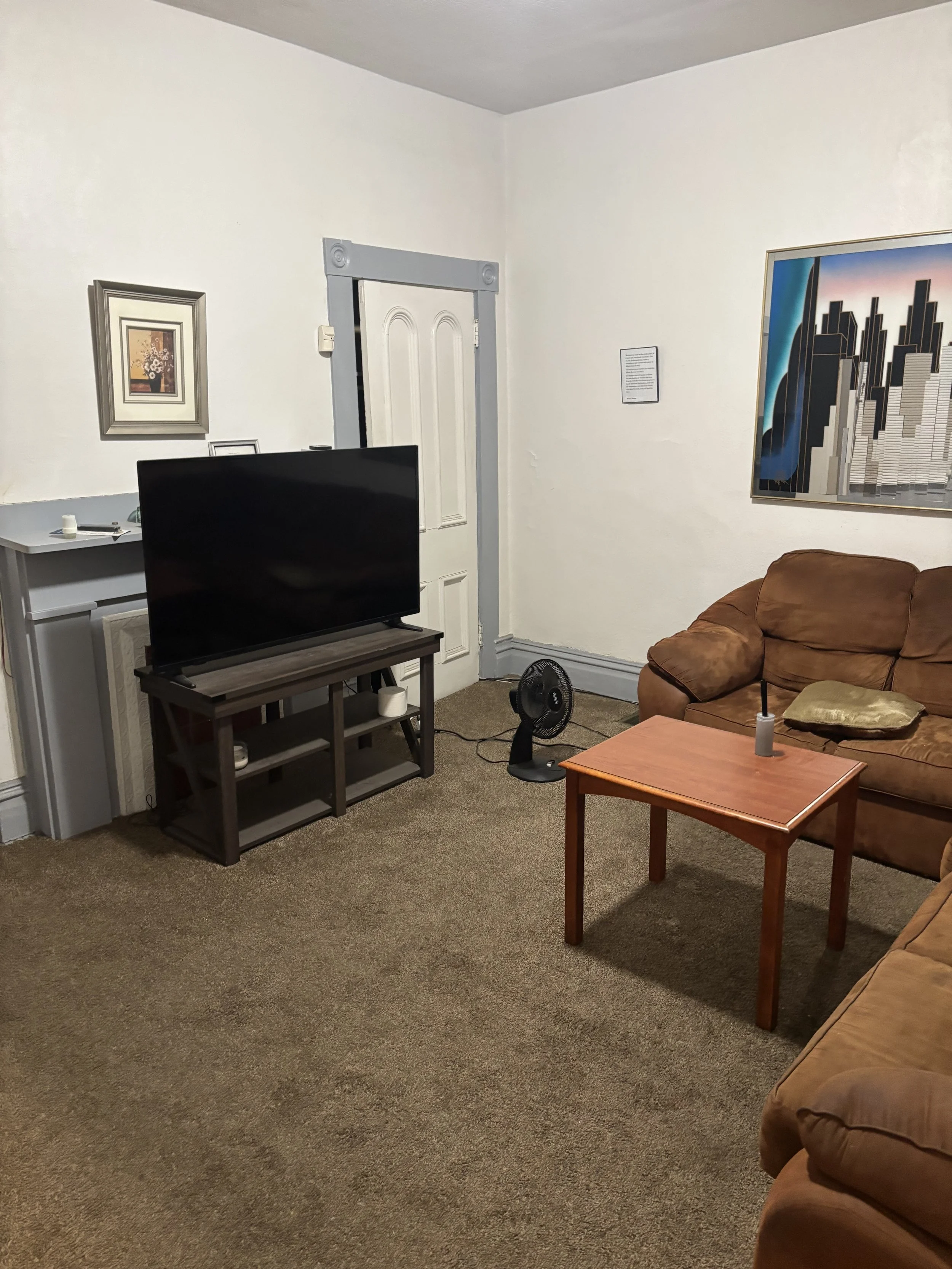 A cozy living room with a brown couch, a small wooden coffee table, a black TV on a stand, a small fan, a framed picture on the wall, a door with decorative trim, and a large framed cityscape art piece.