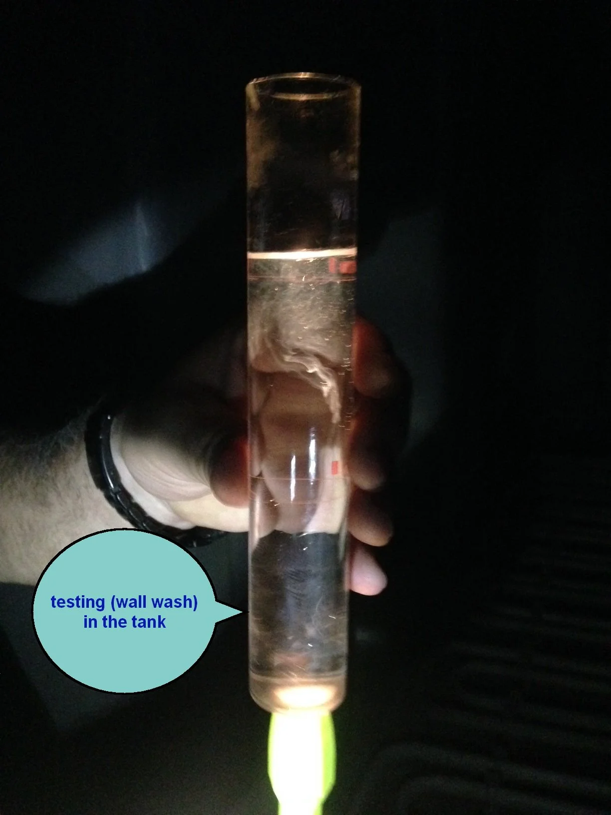 Test tube filled with water in a person's hand, with a green cap at the bottom, used for testing wall wash in a tank, visible in a dark setting.
