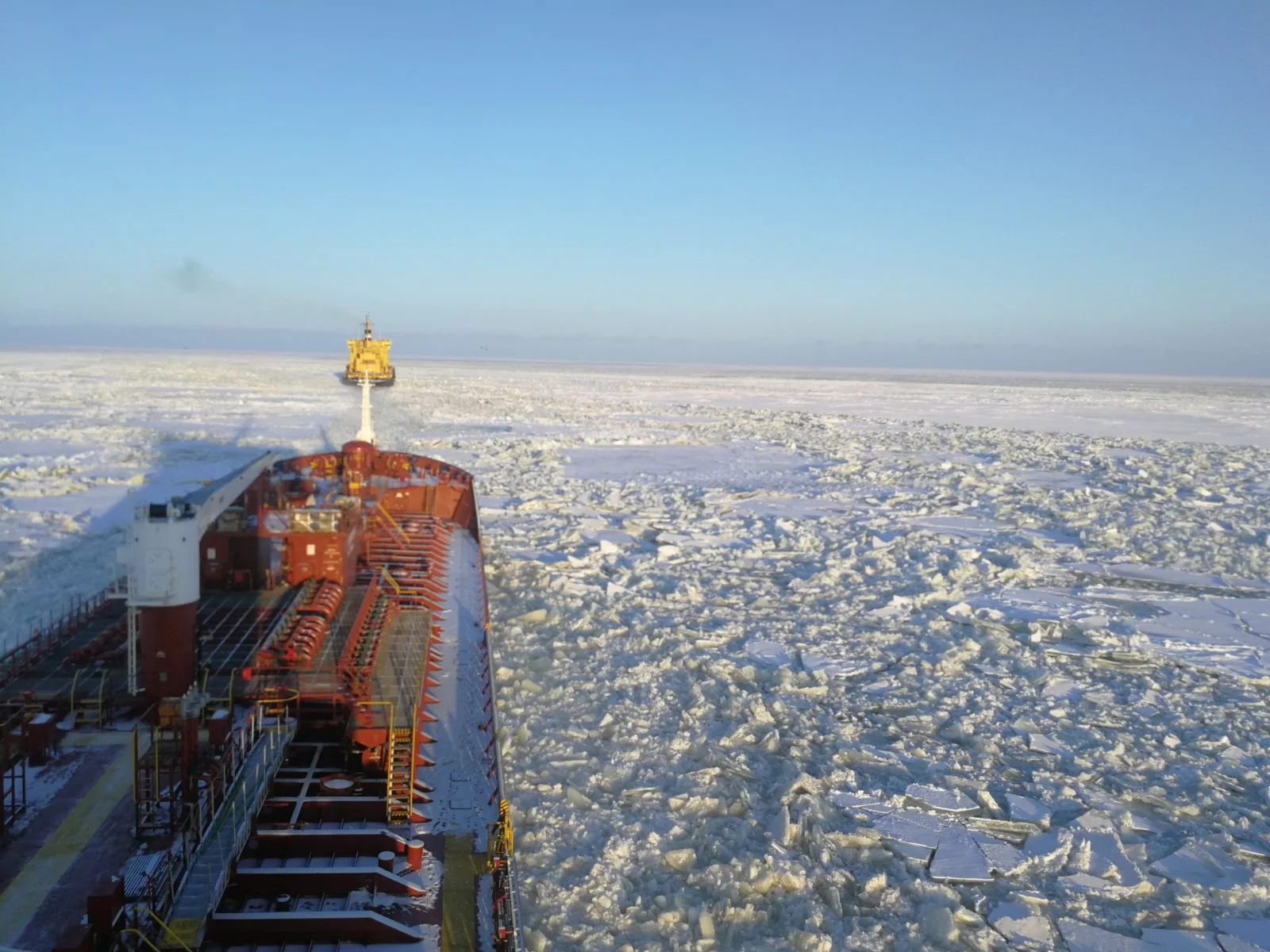 View from a ship navigating through icy waters, with a large icebreaker ship ahead breaking the ice, under a clear blue sky.