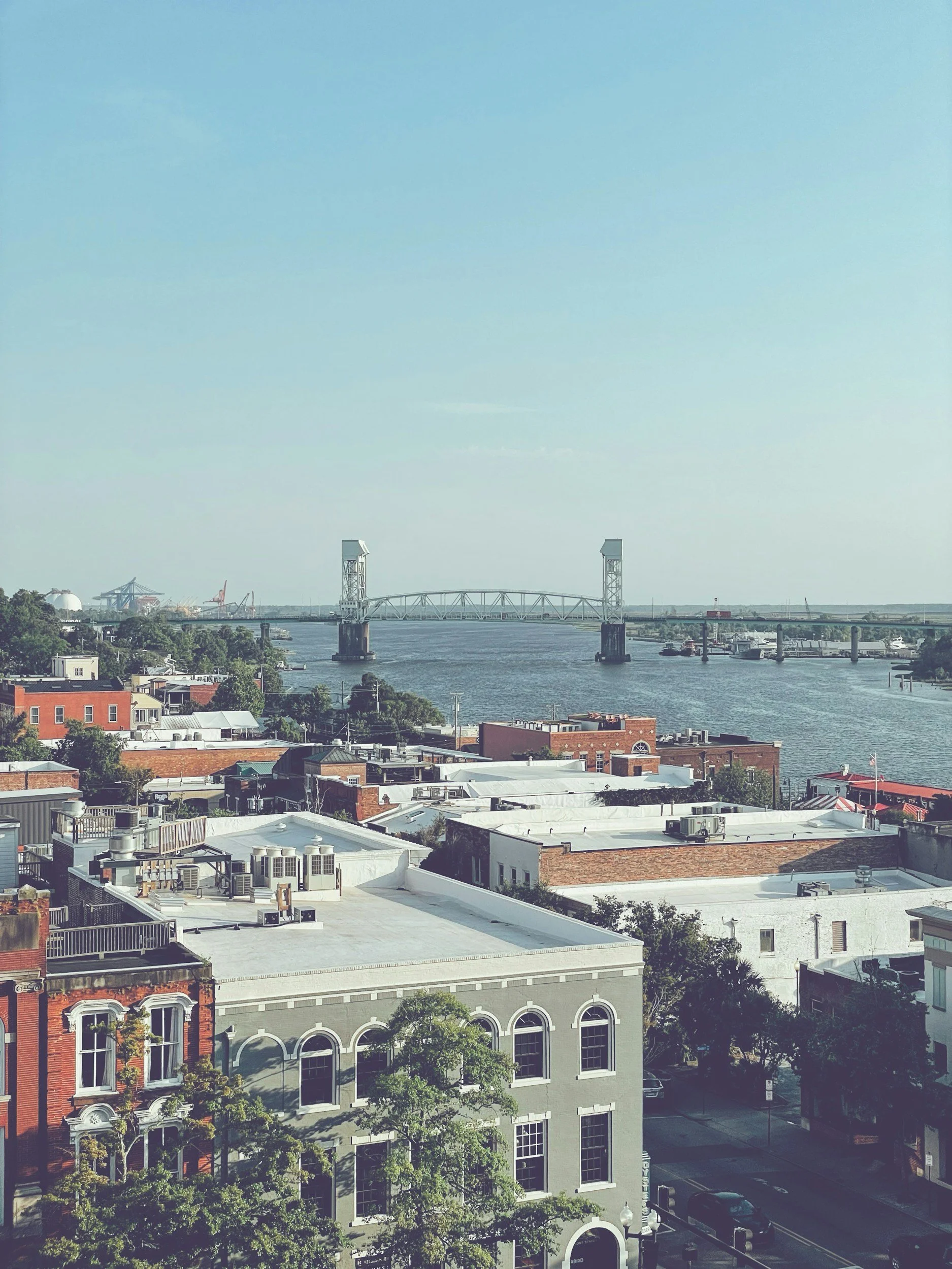 View of a cityscape with rooftops of buildings, a river with boats, and a bridge in the distance under a clear blue sky.