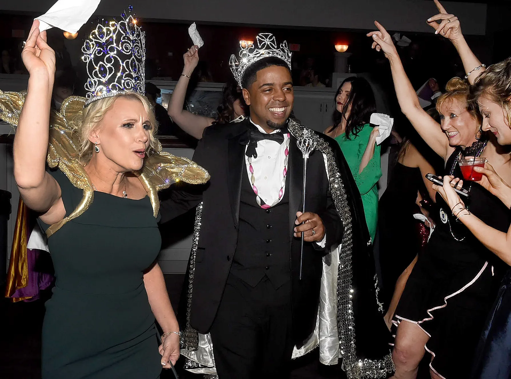 People celebrating at a party, wearing crowns and fancy costumes, with some raising their hands and holding drinks.