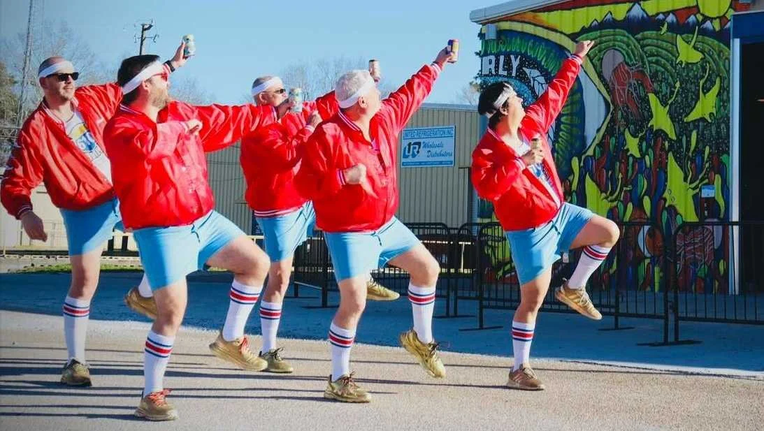 Six men dressed in red jackets, light blue shorts, and striped knee-high socks are dancing together outdoors while holding drinks, with a colorful mural in the background.