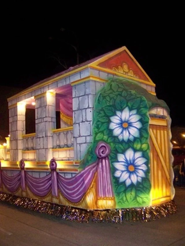 A decorated float resembling a palace with stone walls, purple drapes, and large white and blue flowers painted on the side, illuminated at night.