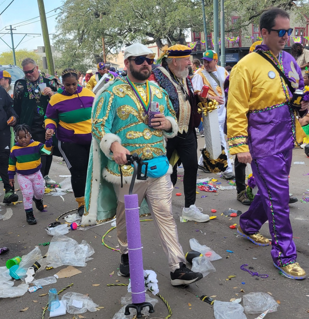 People celebrating Mardi Gras parade wearing colorful costumes, with beads and decorations, on a street with scattered trash and confetti.