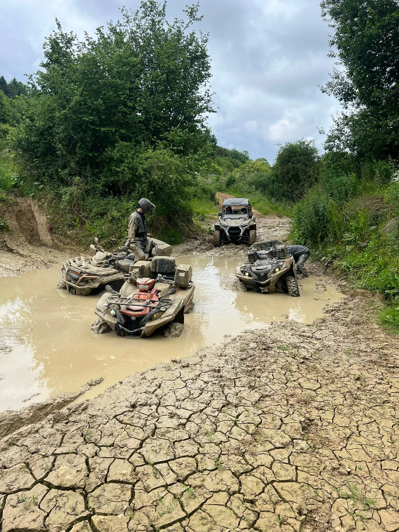 Group of people riding all-terrain vehicles through a muddy water crossing on a rugged outdoor trail with trees and cloudy sky.