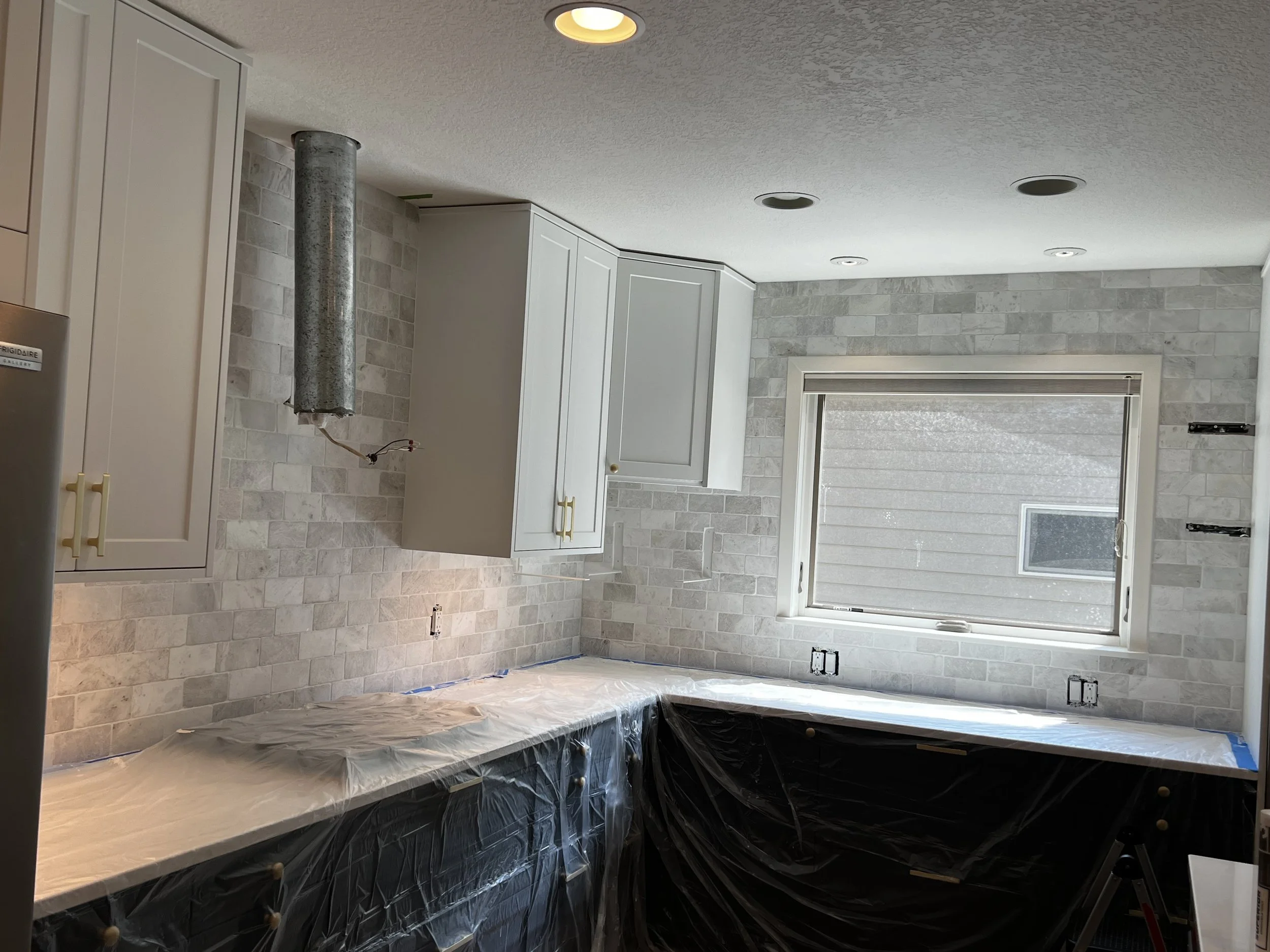 Kitchen under renovation with white cabinets, gray tiled backsplash, plastic-covered countertops, and a window on the back wall.