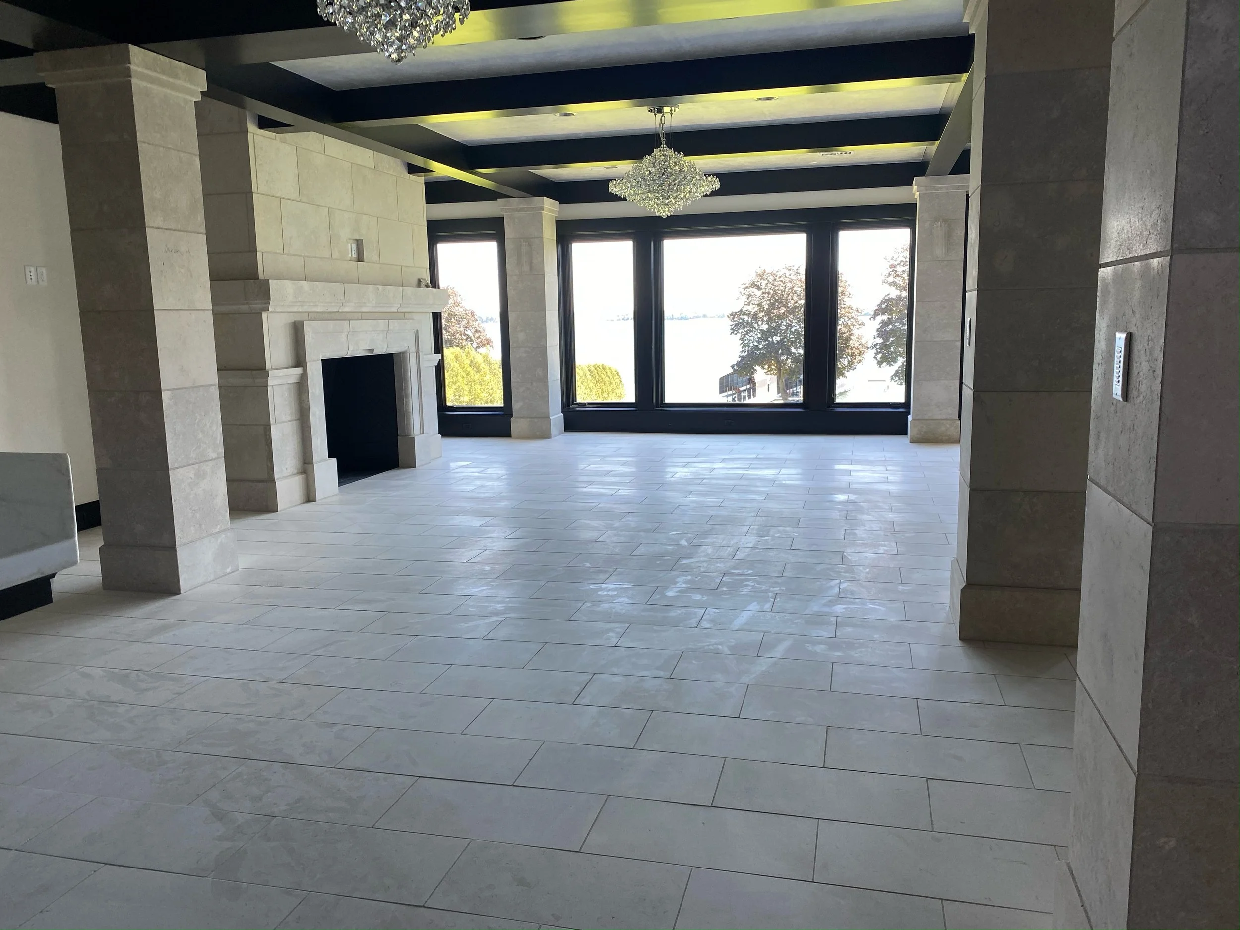 Empty modern living room with large windows, a fireplace, chandeliers, and tiled floor.