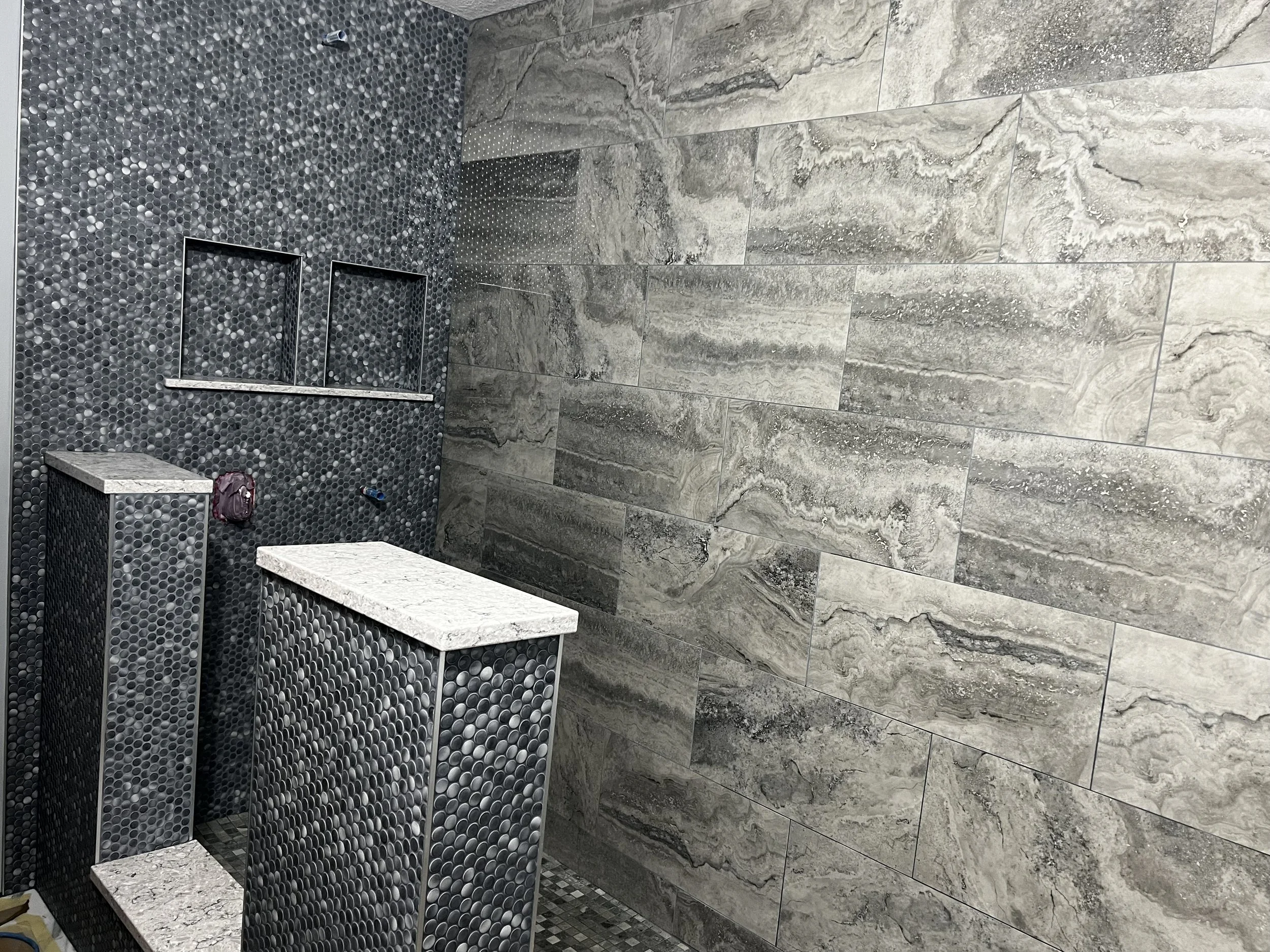 Bathroom with gray stone tiles on the wall and floor, dark gray pebble mosaic on the wall, and three metallic shower niches. There are two rectangular blocks with a gray and white stone finish and a small red bag on the floor.