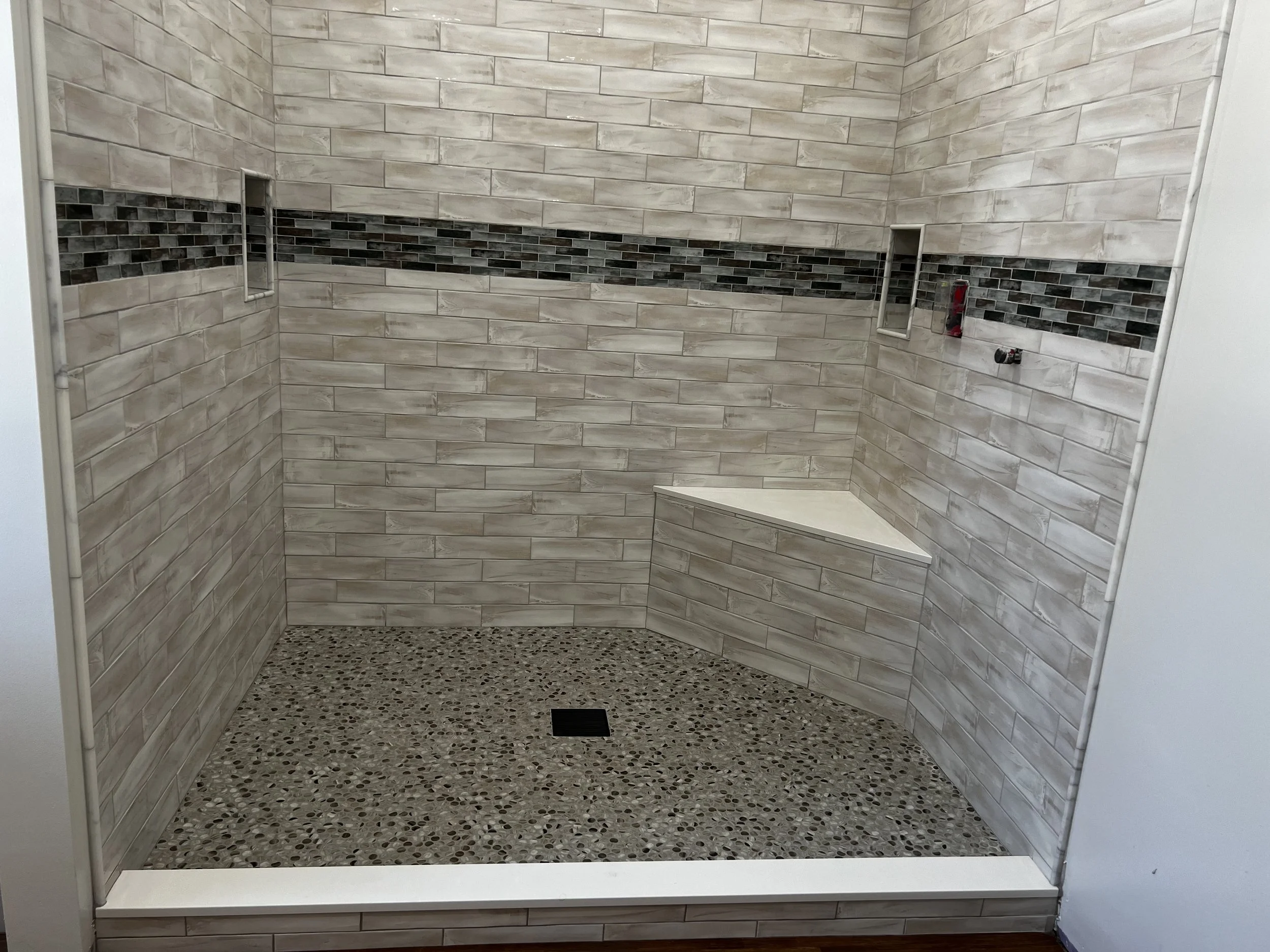 Empty walk-in shower with beige and gray tile walls, a dark mosaic accent strip, a built-in corner seat, and a drain in the pebble tile floor.