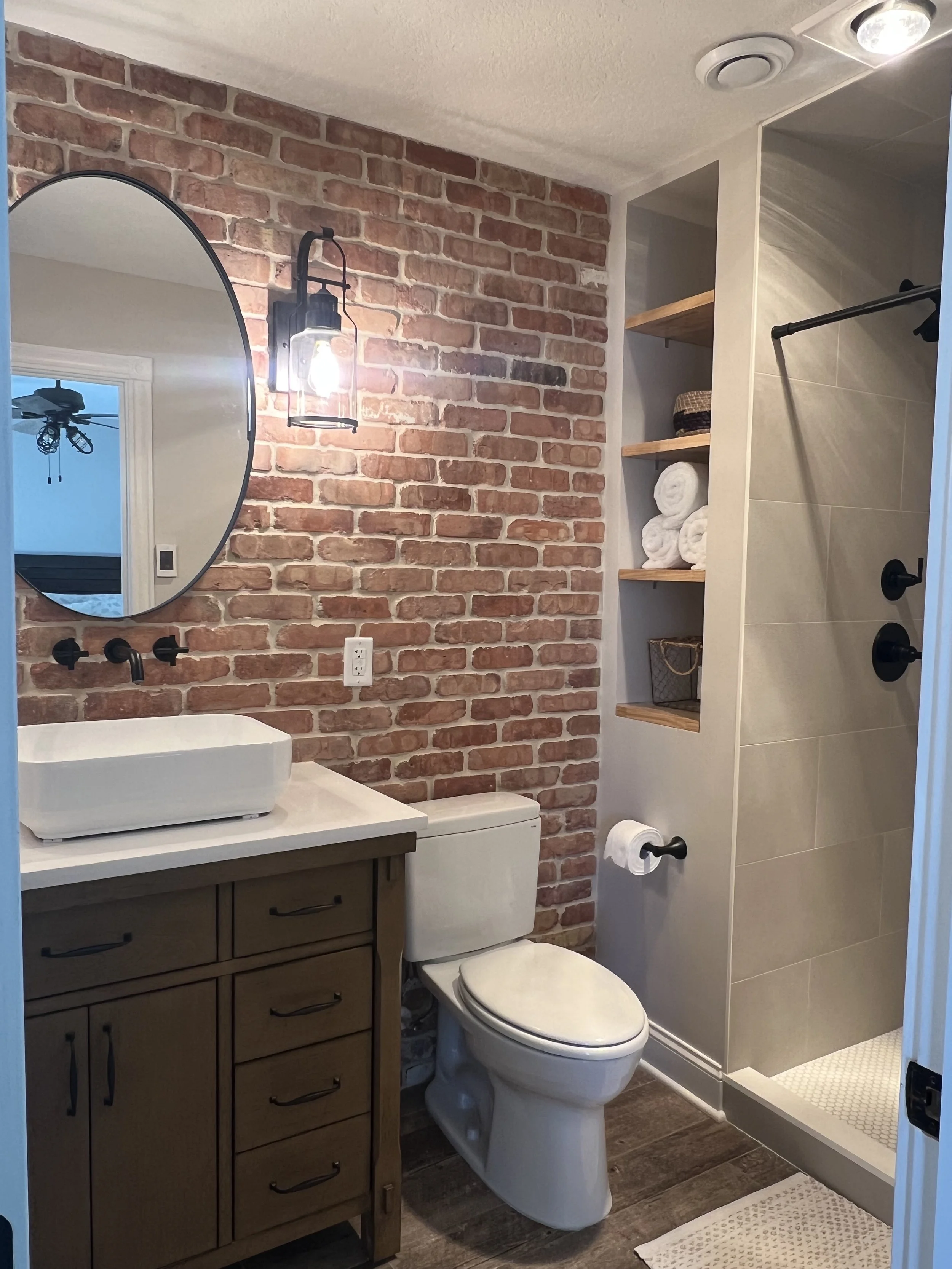 A modern bathroom with exposed brick wall, wooden vanity with a vessel sink, round mirror, wall-mounted black faucet, wall sconce, toilet, open shelving with folded towels, a woven basket, and a shower area with beige tiles and black fixtures.