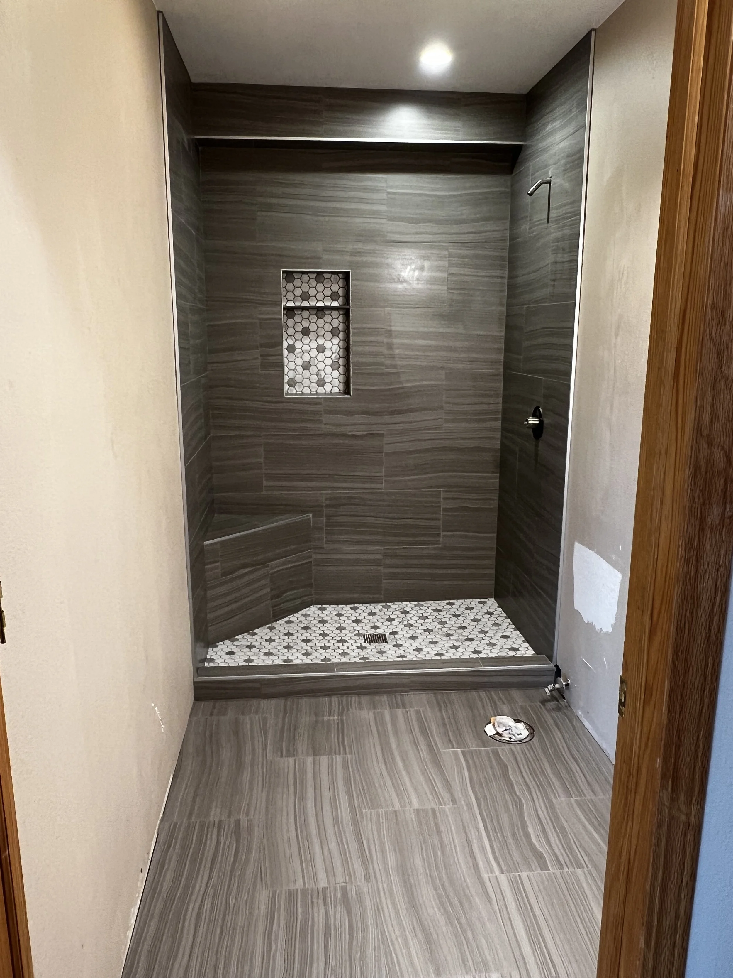 An empty walk-in shower with gray tiles on the walls and floor, a built-in niche with hexagonal tiles, and a small bench in the corner. There is a recessed ceiling light and plumbing fixtures, with some construction or finishing work visible on the a