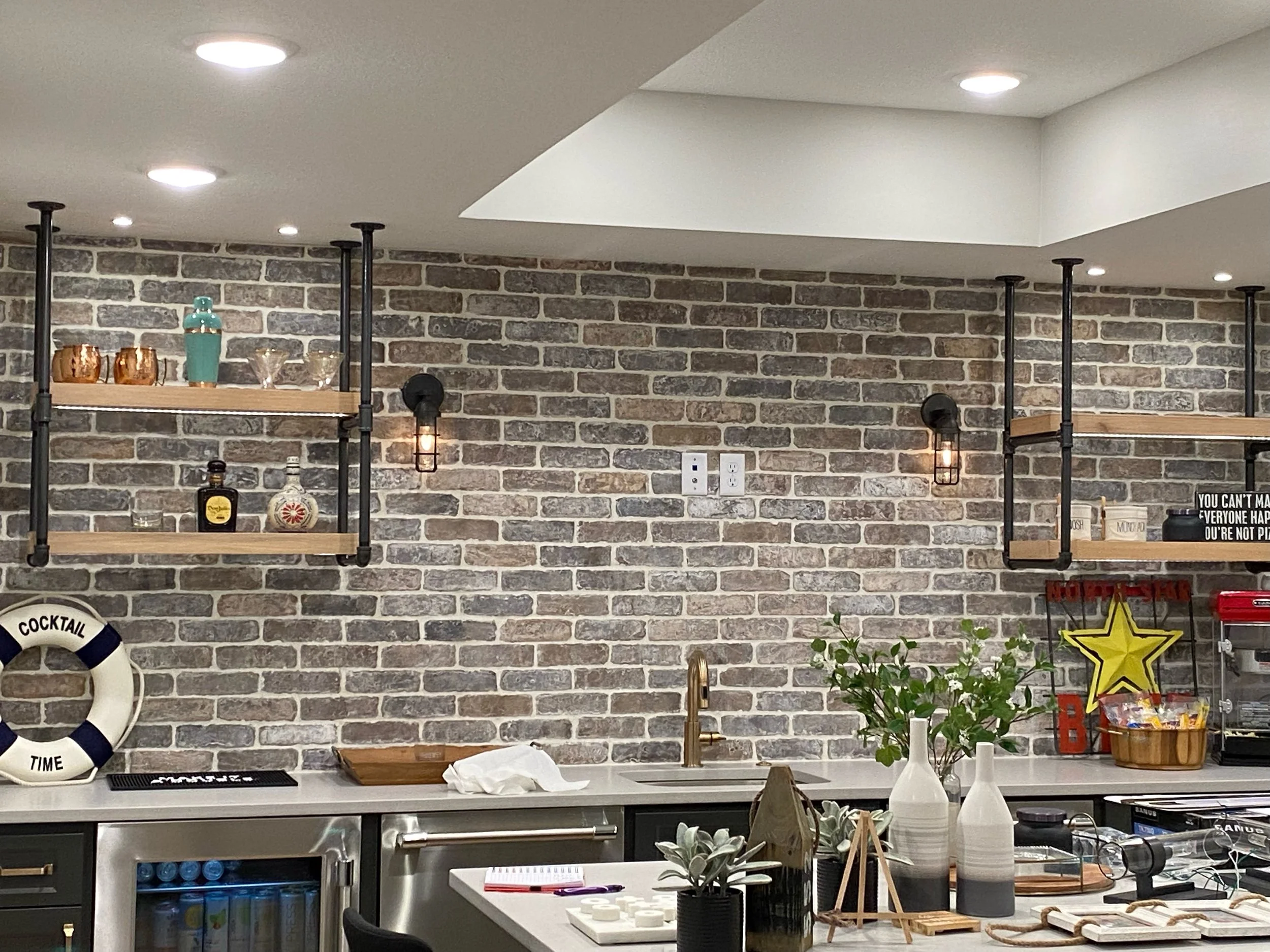 Modern kitchen with brick accent wall, floating shelves, and decorative items including bottles, glasses, and a life preserver. Countertop with plants, vases, and various kitchen accessories.
