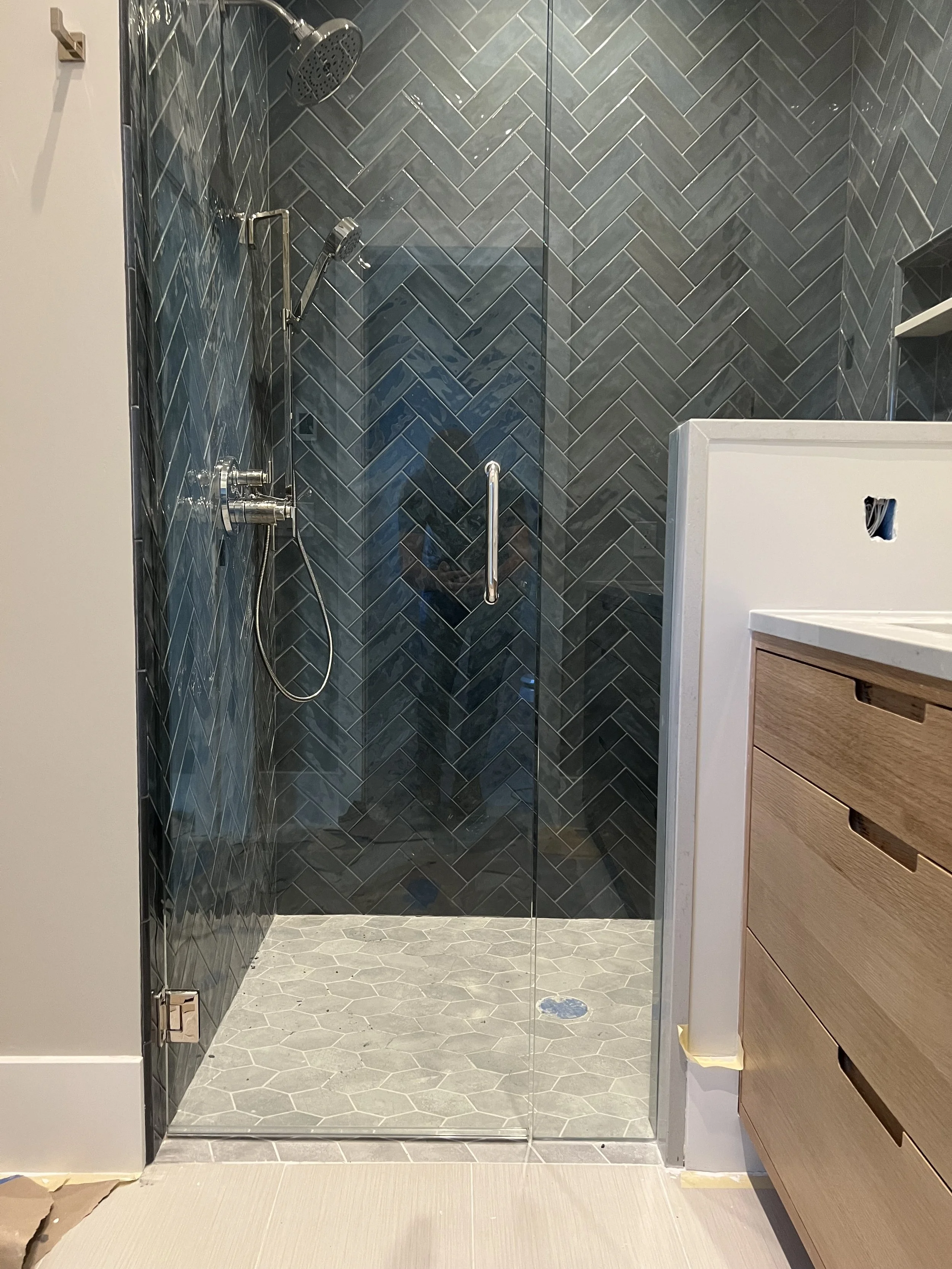 Modern walk-in shower with dark chevron tile walls, light hexagon tile floor, and a glass door, adjacent to a wooden vanity cabinet.