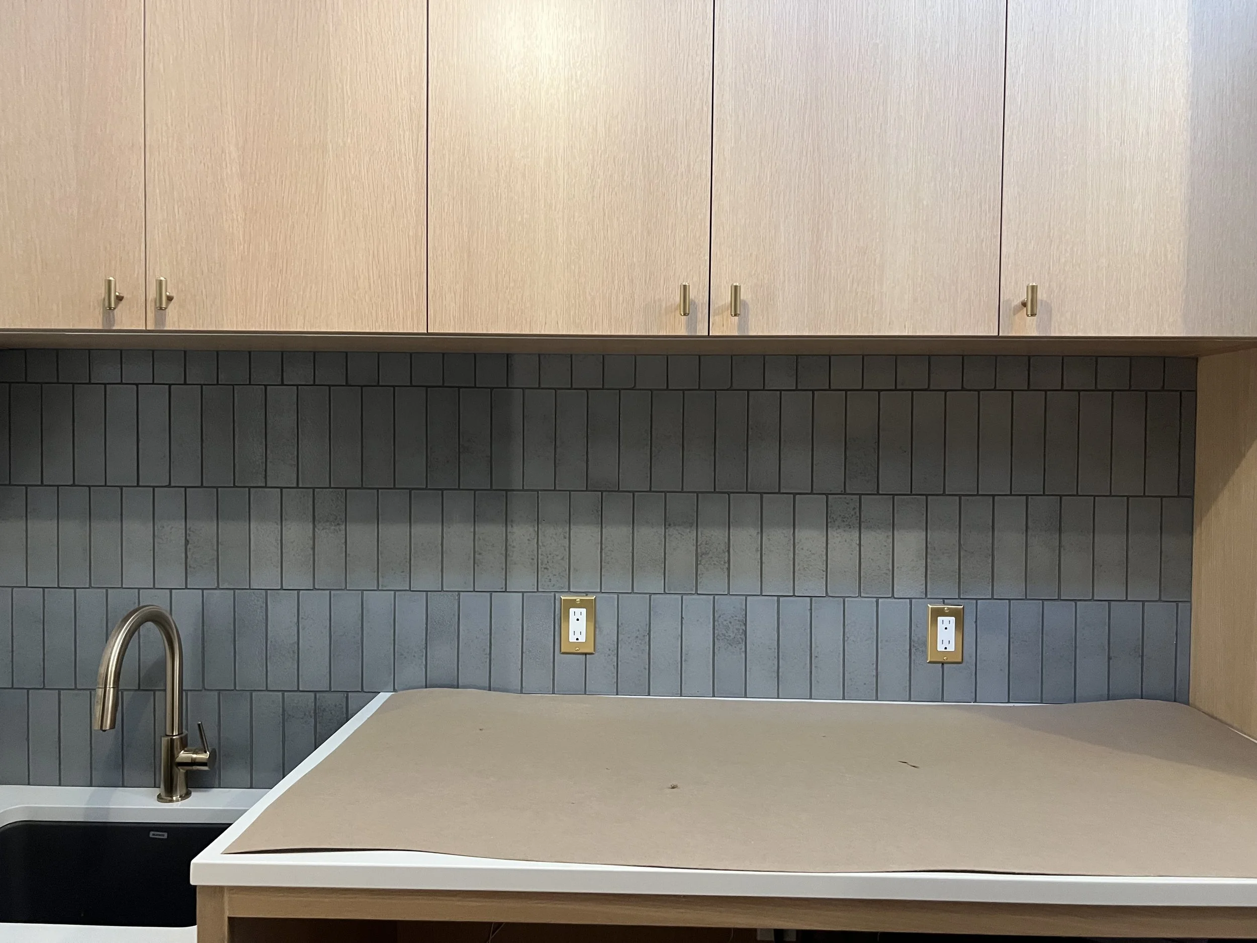 Kitchen countertop with beige covering, silver faucet, dark backsplash tiles, two electrical outlets, beige wall cabinets at the top.