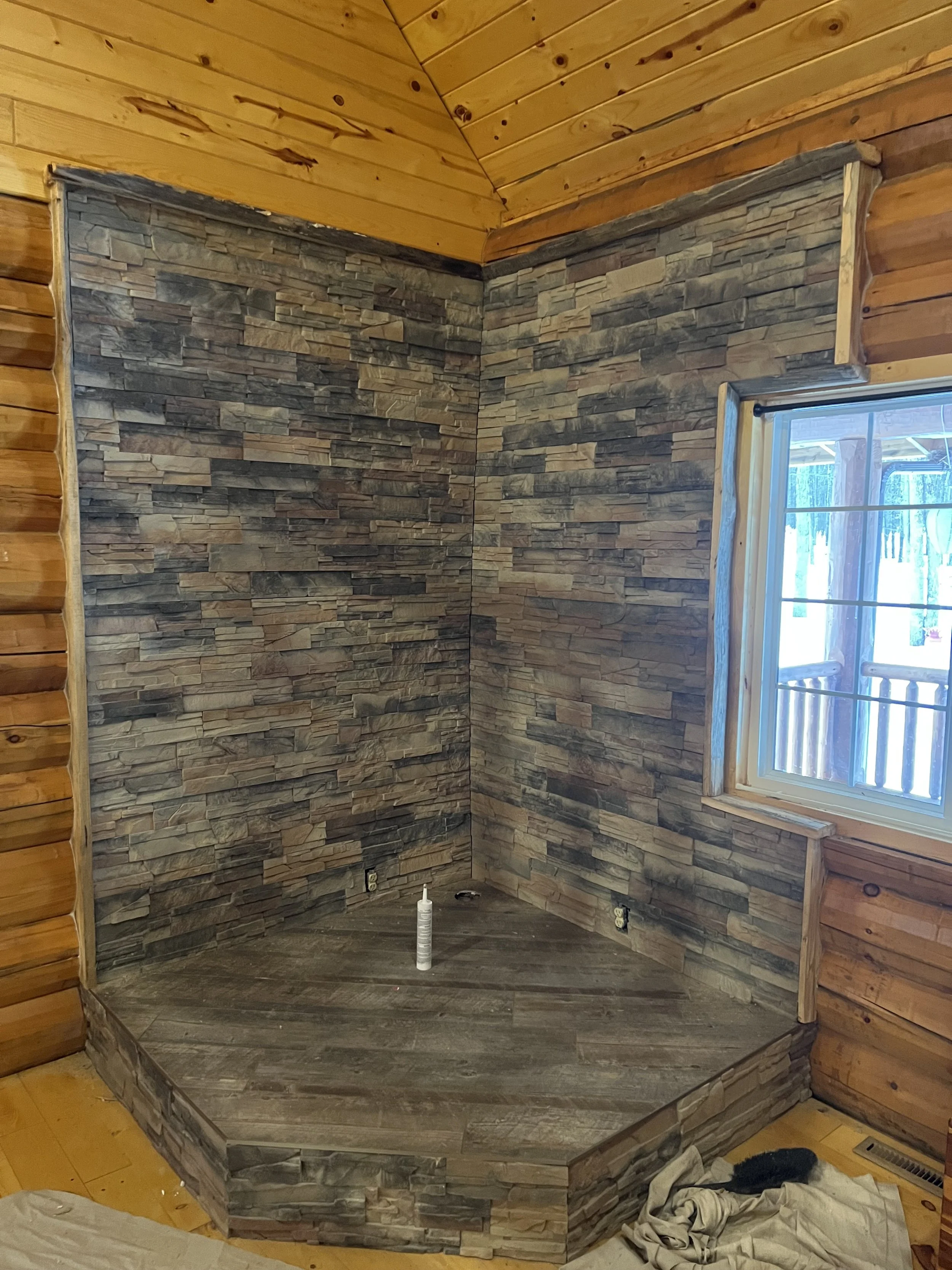 Corner of a room under construction with stone and wood finishing, a window with a wooden frame, and a towel and tool on the floor.