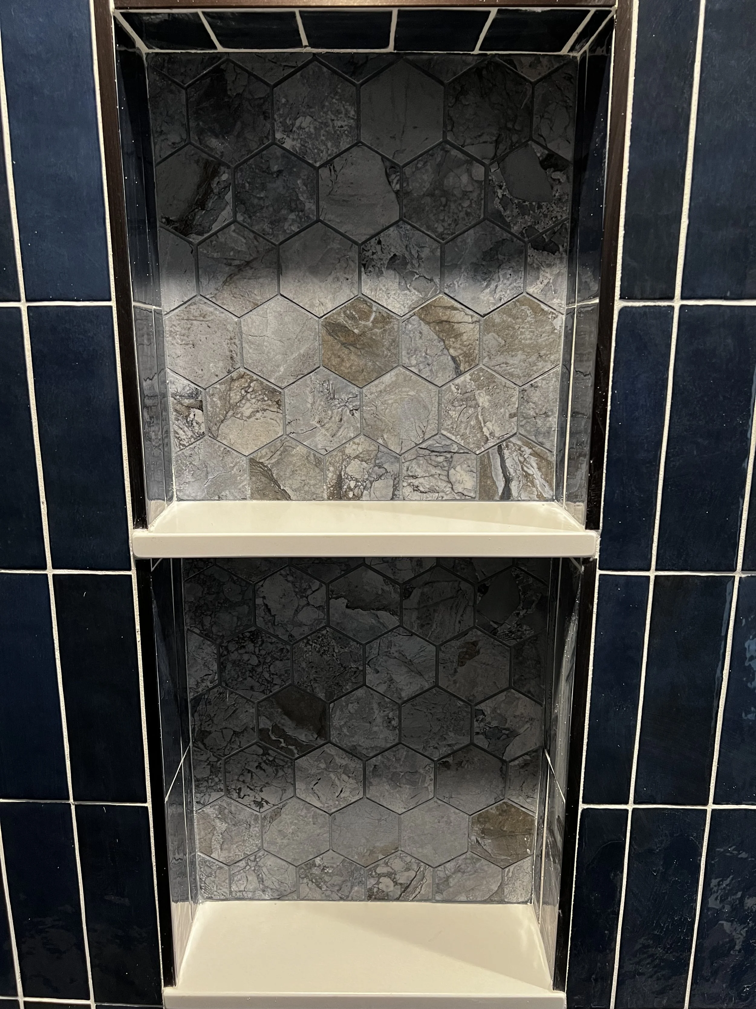 A built-in shower niche with two shelves, featuring black hexagon tiles on the back wall with gray and beige marbled tile accents, surrounded by dark blue tiled walls with white grout.