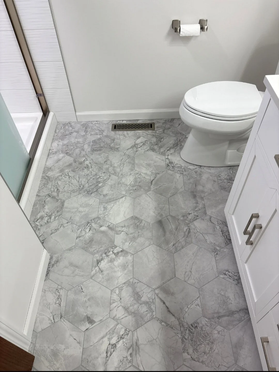 Bathroom with marble hexagonal tile flooring, a white toilet, a white vanity, a toilet paper holder with a roll of toilet paper, and a vent on the floor.