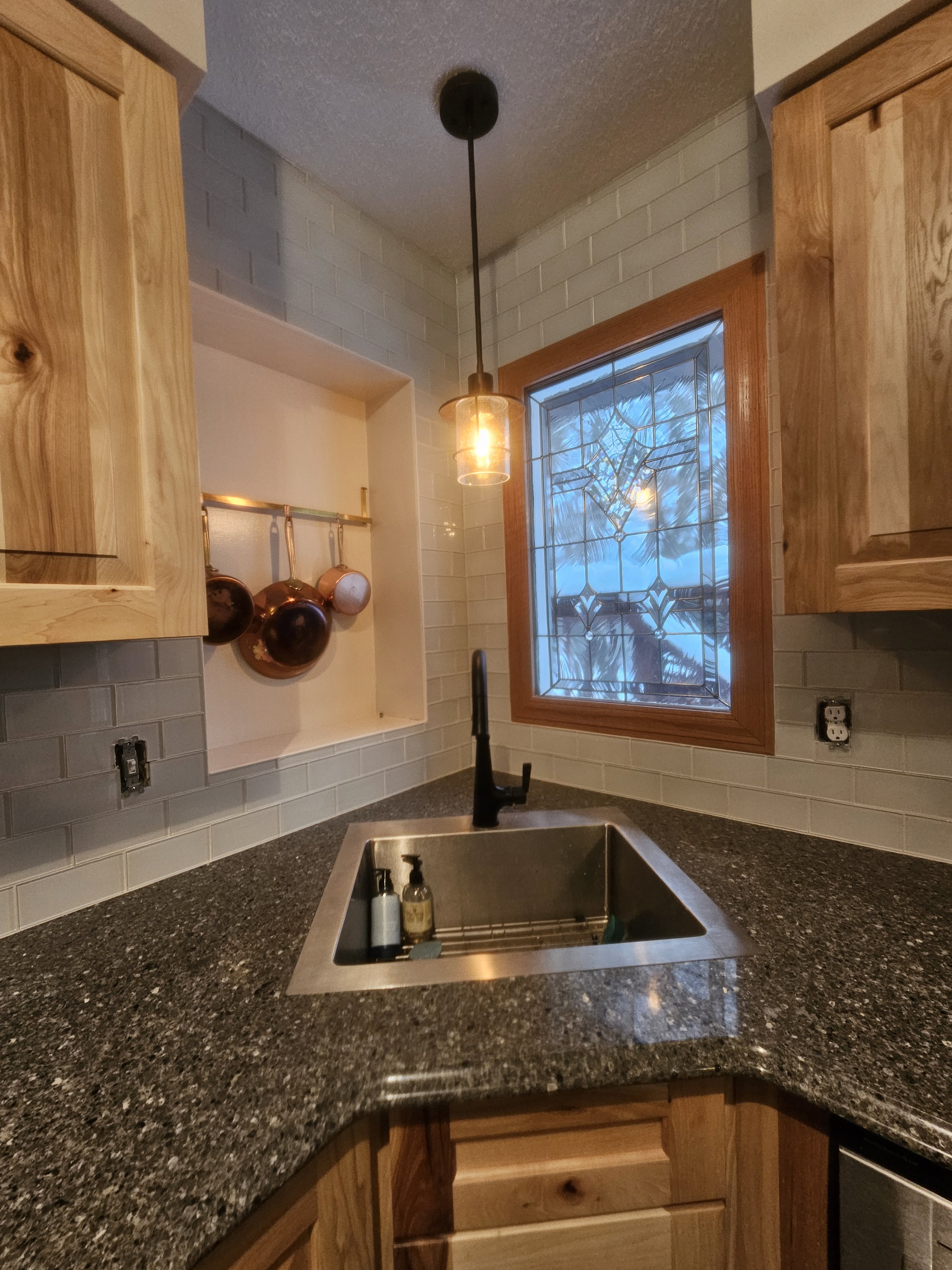 Kitchen corner with a granite countertop, stainless steel sink, black faucet, wooden cabinets, a window with stained glass, a hanging light fixture, and copper pots hanging on the wall.
