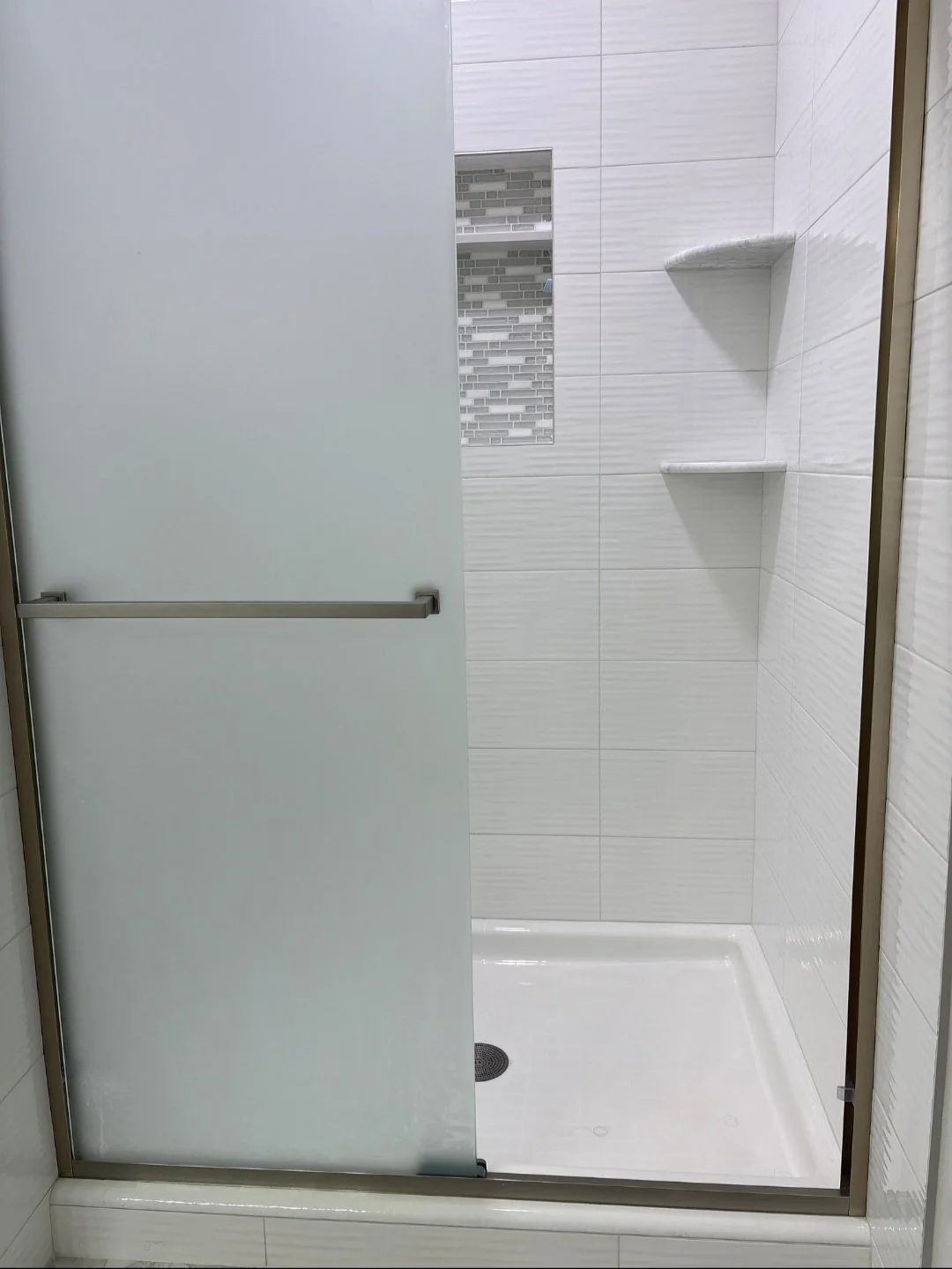 A modern shower stall with white tiled walls, a small built-in shelf, a frosted glass door, and a shower drain on the white shower base.
