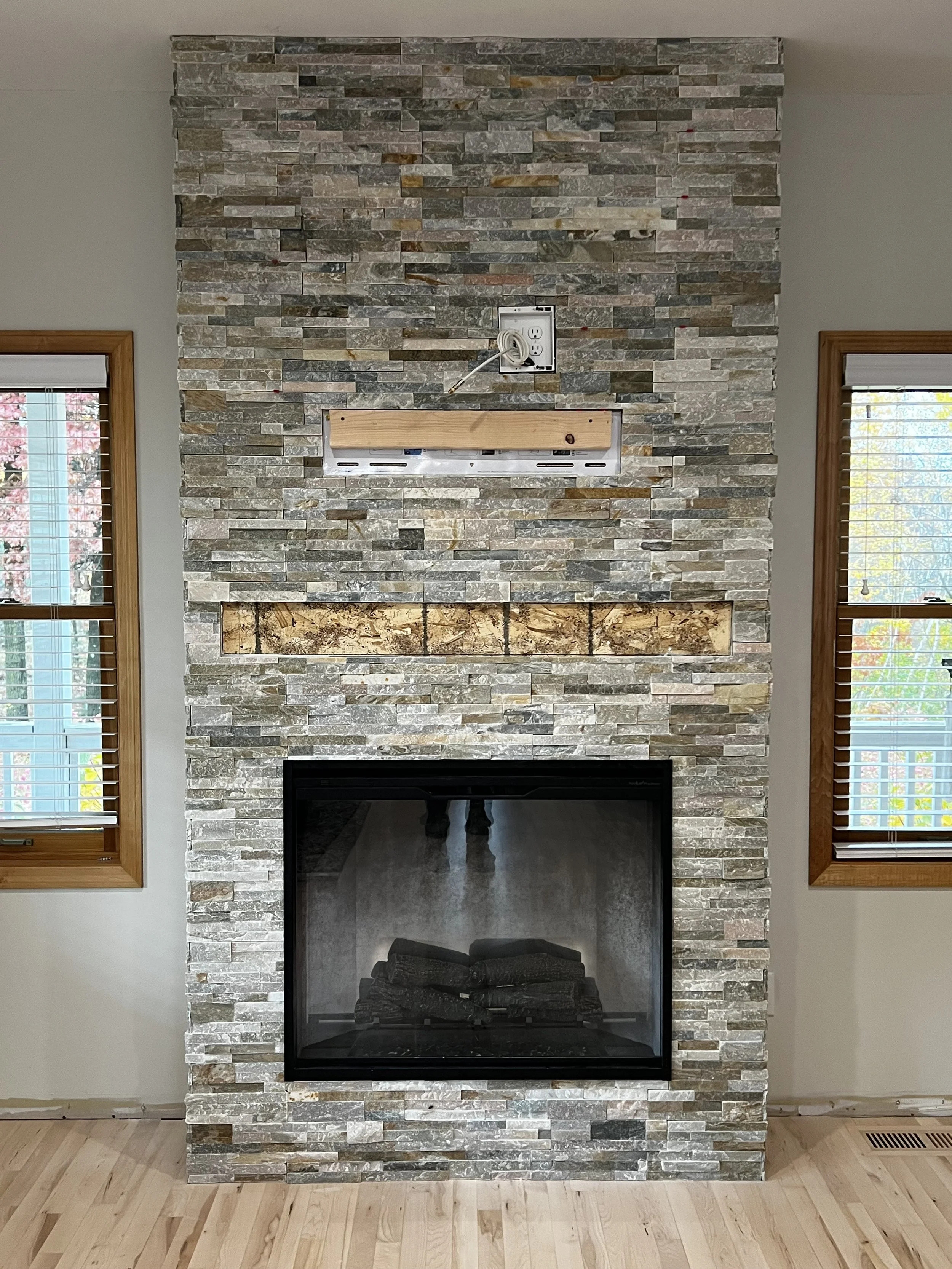 Stone fireplace with a black glass front, surrounded by gray and tan stacked stone, two wooden window frames with blinds on either side, and unfinished mantel area with electrical outlet and hanging cord.