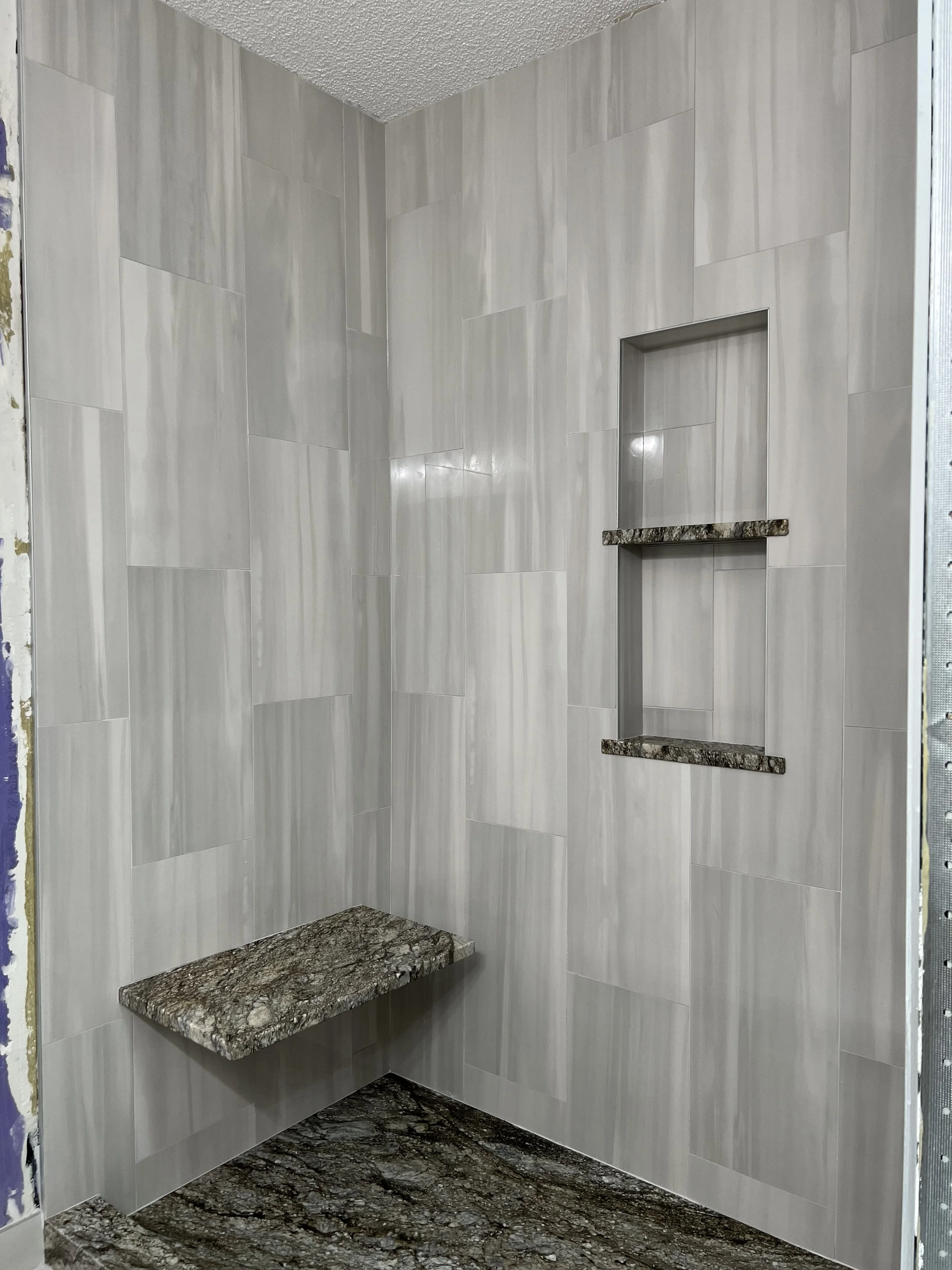 Newly tiled shower with light gray rectangular wall tiles, a built-in niche with granite shelves, and a granite bench.