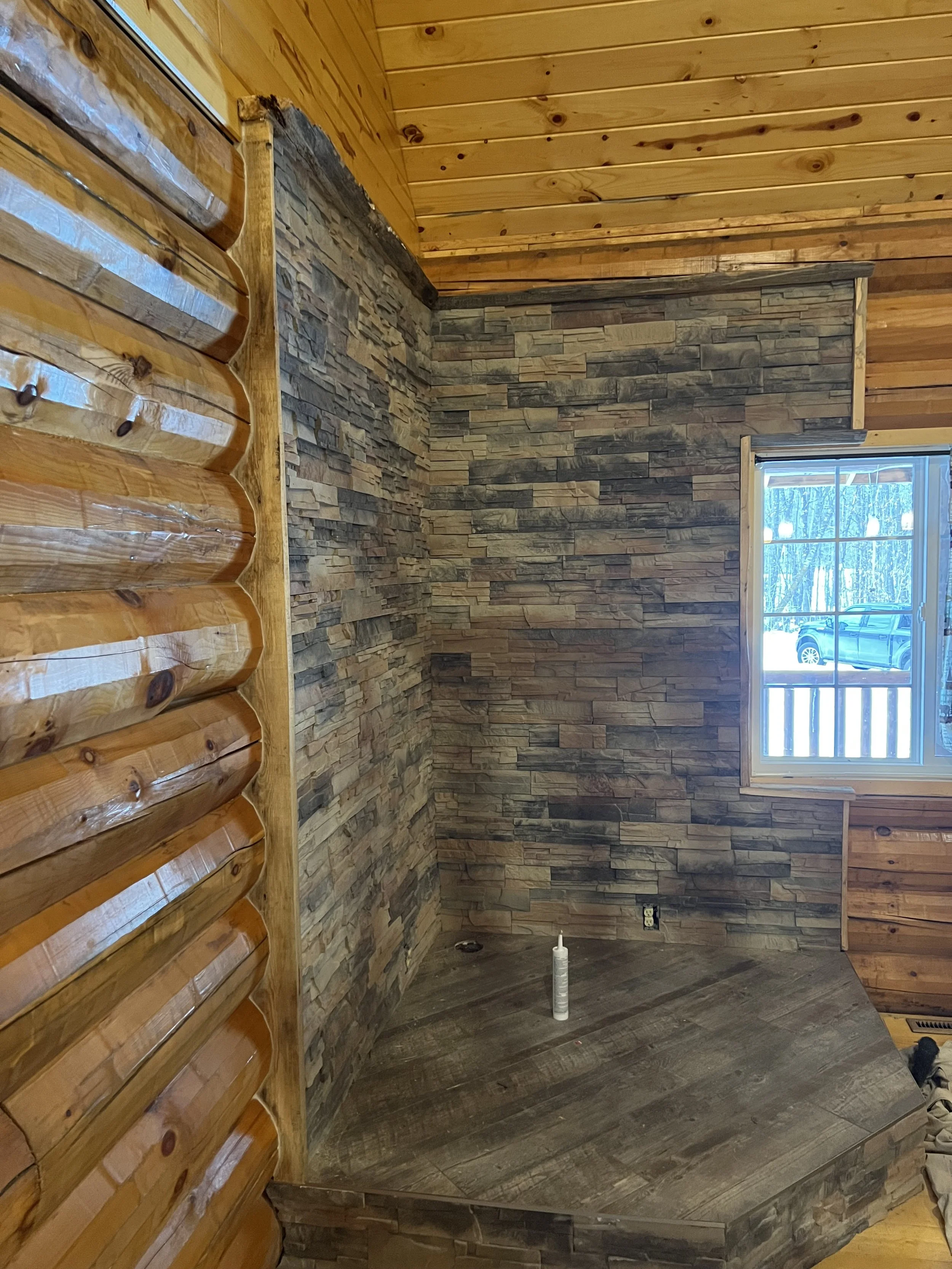 Interior of a room with wooden log walls, a brick accent wall, wooden ceiling, and a window revealing an outdoor scene with a vehicle.